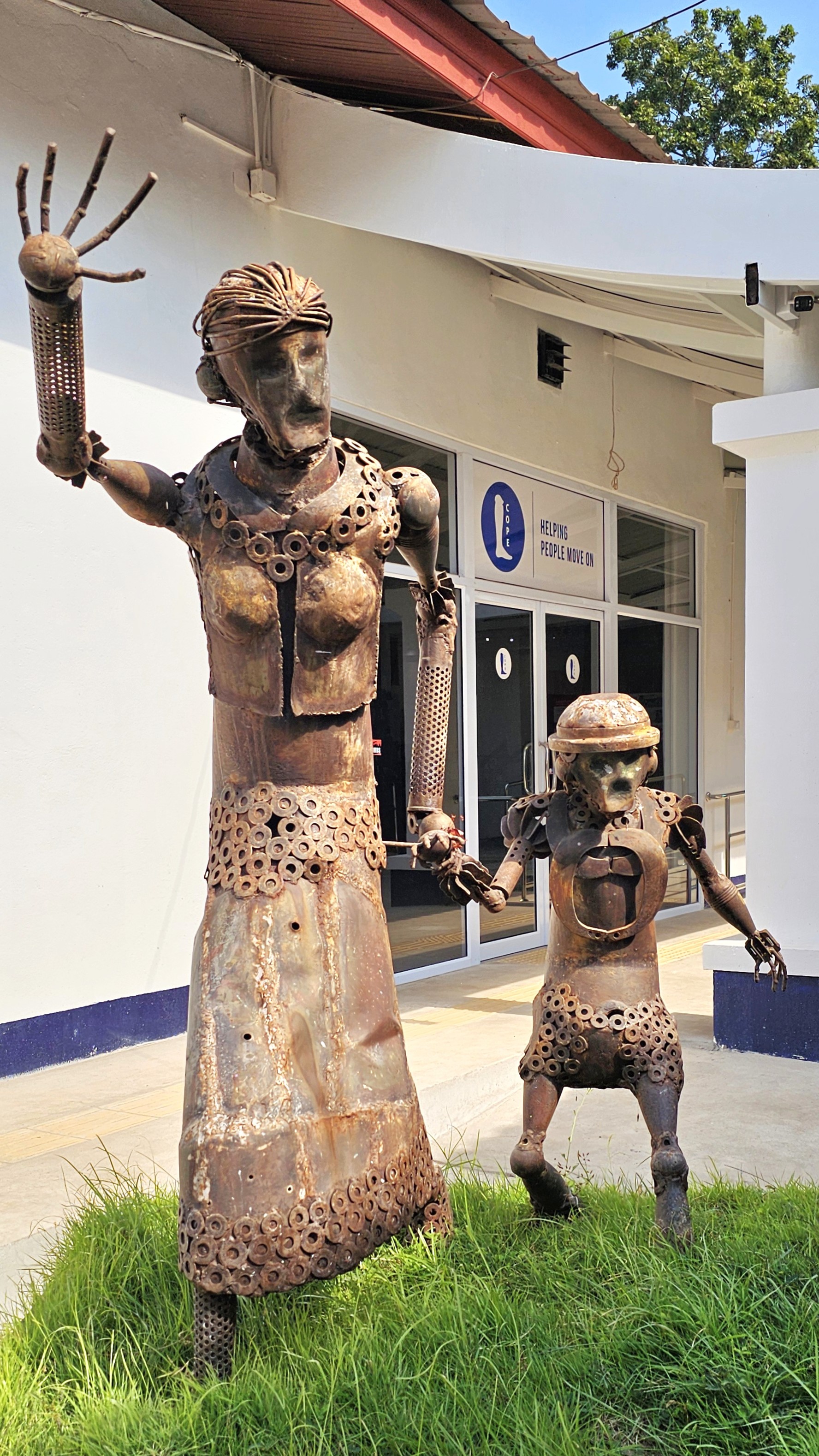 Sculptures of a woman and a child made from metal scraps, standing on a patch of grass outside a building with a sign that reads 'Helping People Move On'.