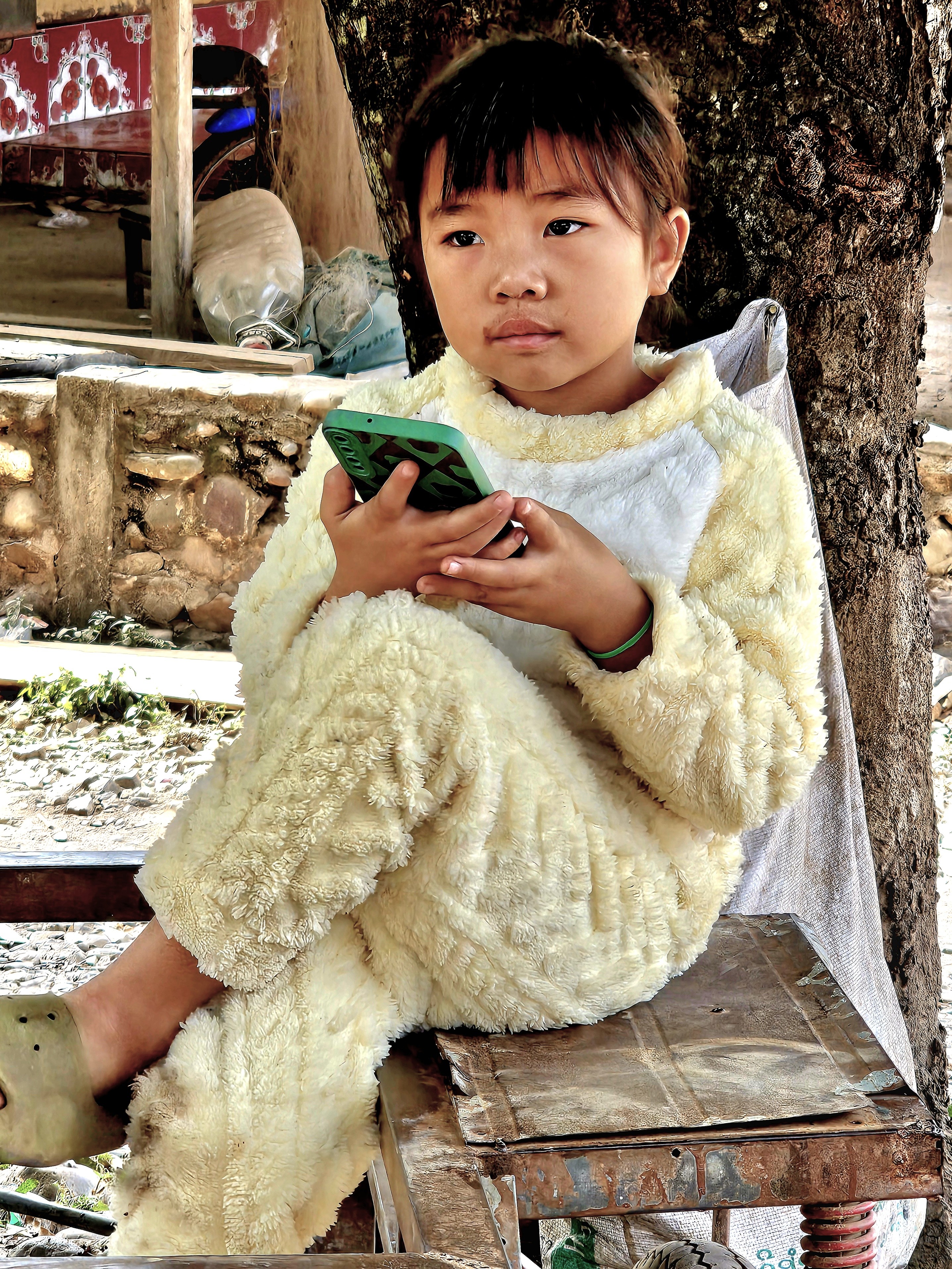 A young child in a fluffy, light-colored outfit thoughtfully holds a smartphone while sitting on a wooden stool next to a tree in a rustic environment.