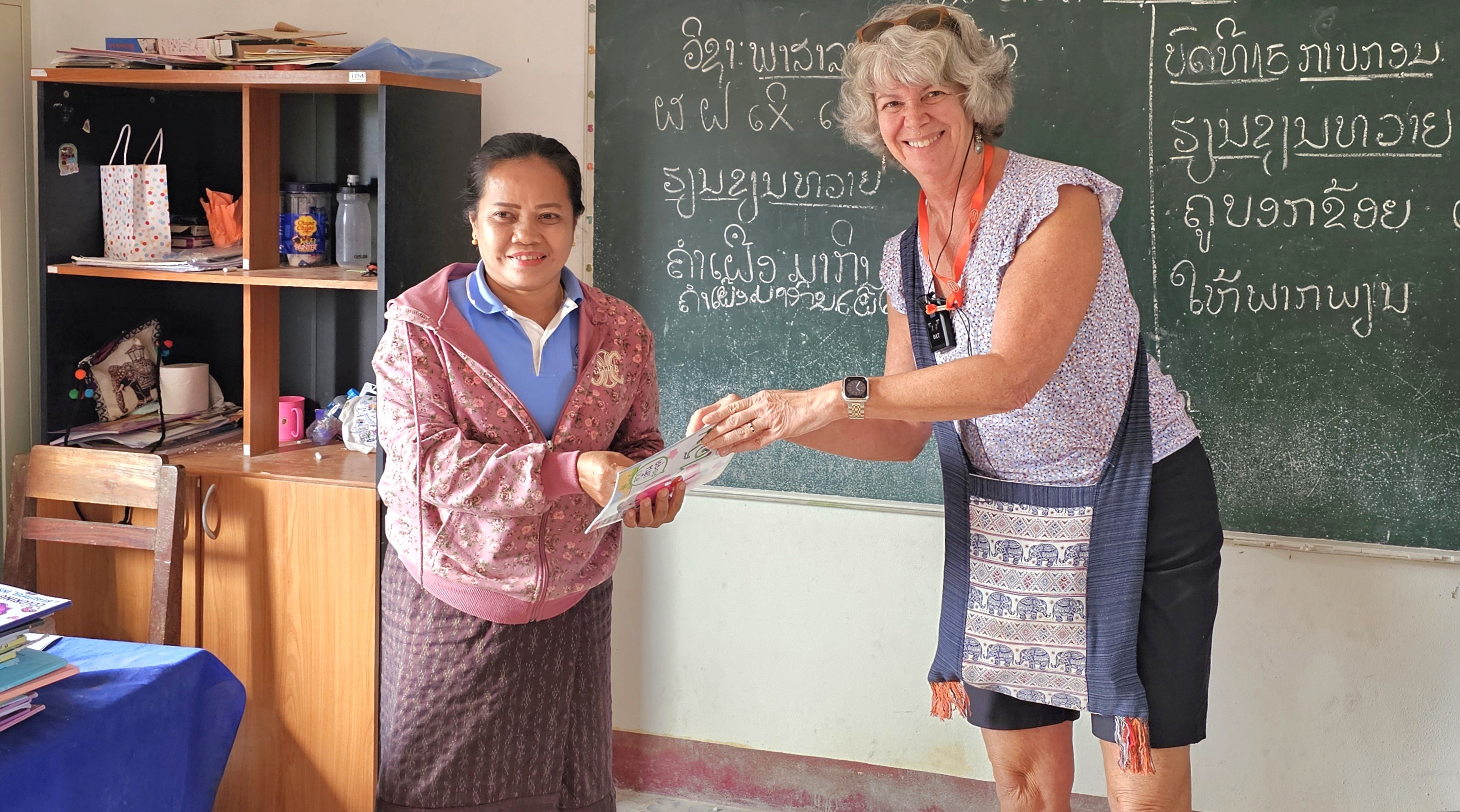 Two women interact in a classroom setting, with one handing a booklet to the other, who is smiling. A chalkboard with writing in Lao is visible in the background along with classroom materials.