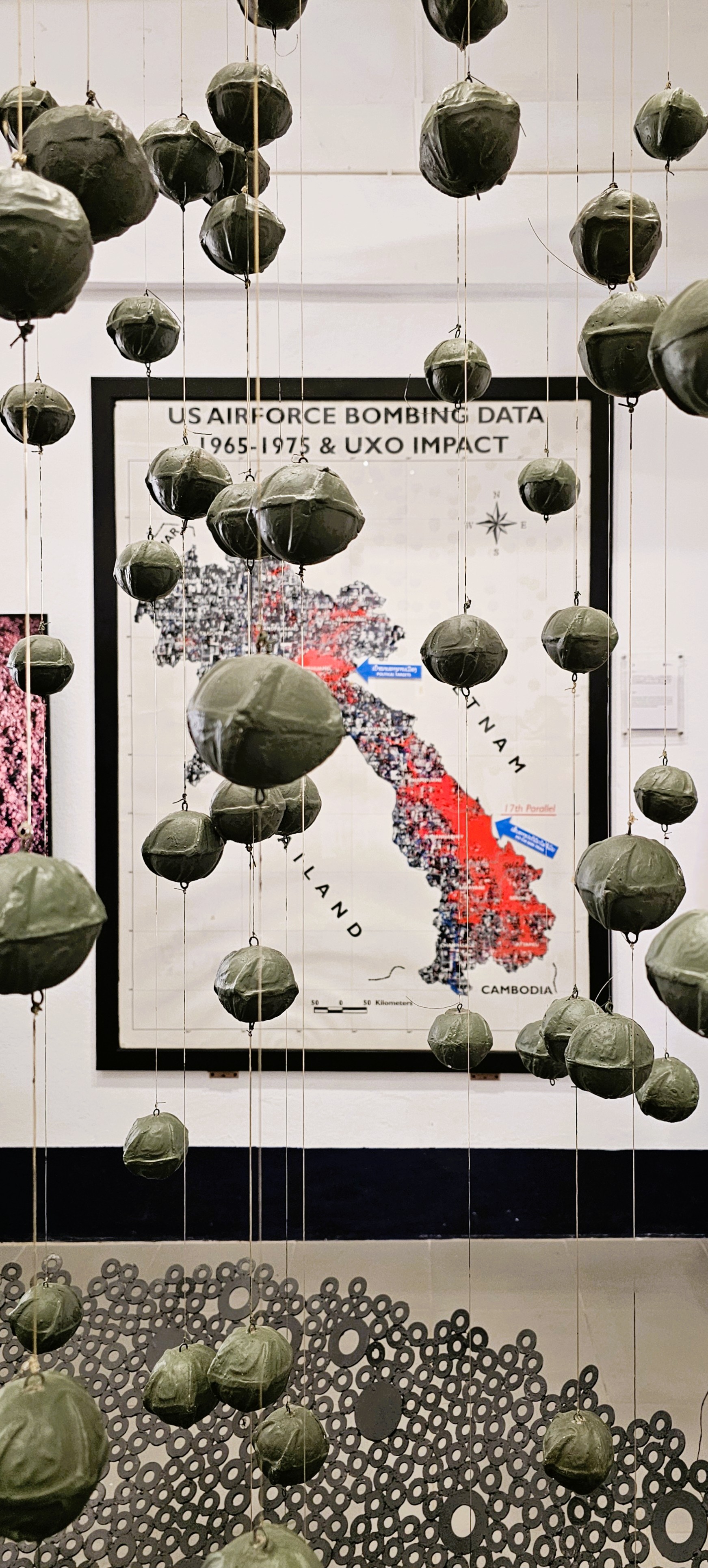 Hanging art installation resembling bombs, with a background featuring a map illustrating US Airforce bombing data and unexploded ordnance impacts in Laos from 1965 to 1975.