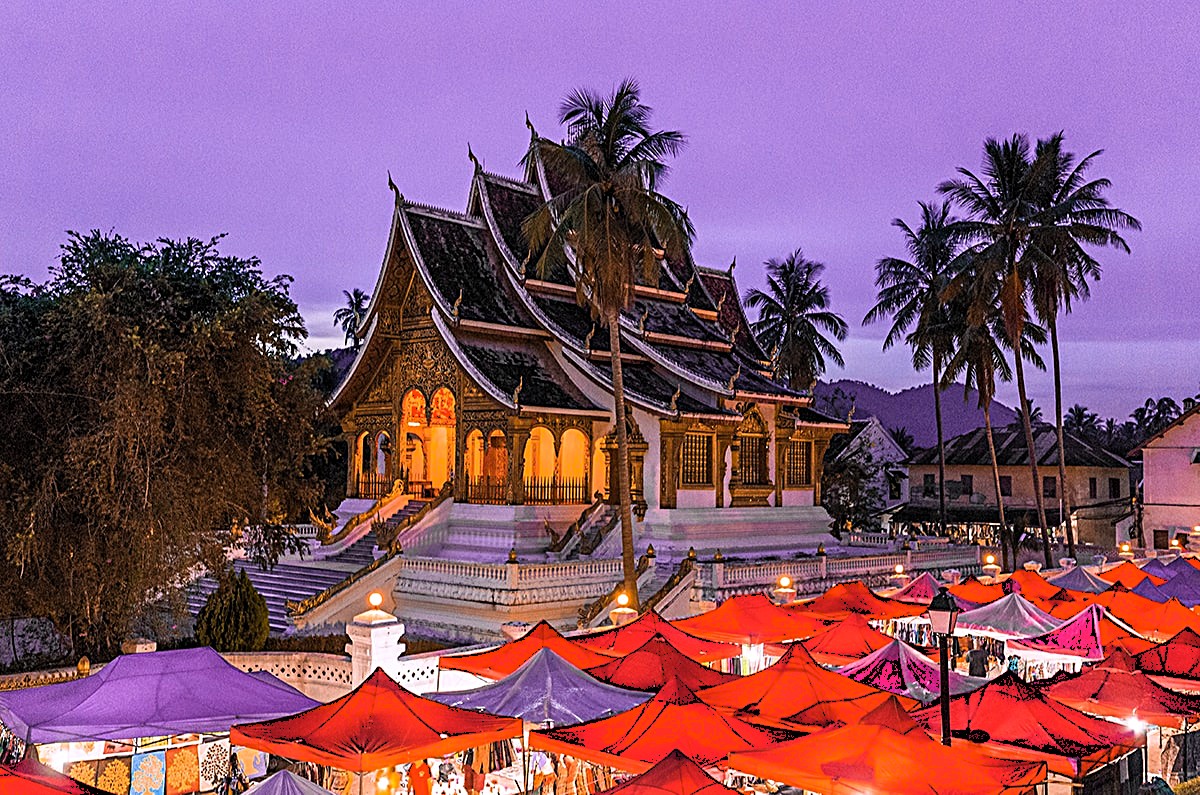 A view of a traditional Lao temple illuminated at dusk, surrounded by colorful market stalls with red canopies in the foreground, and palm trees against a twilight sky.