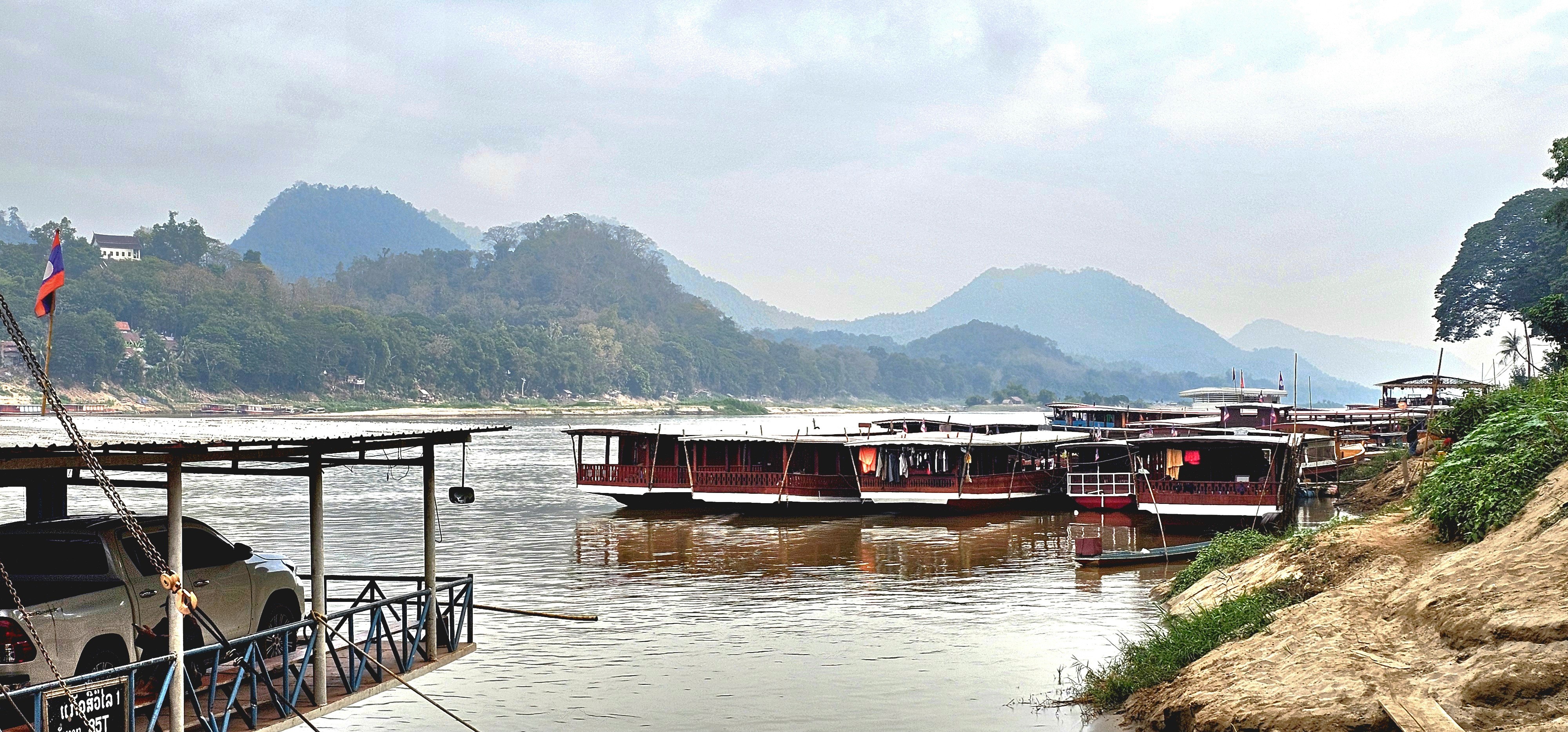Scenic view of the Mekong River with traditional wooden boats moored at a riverside pier, surrounded by lush green hills under a cloudy sky.