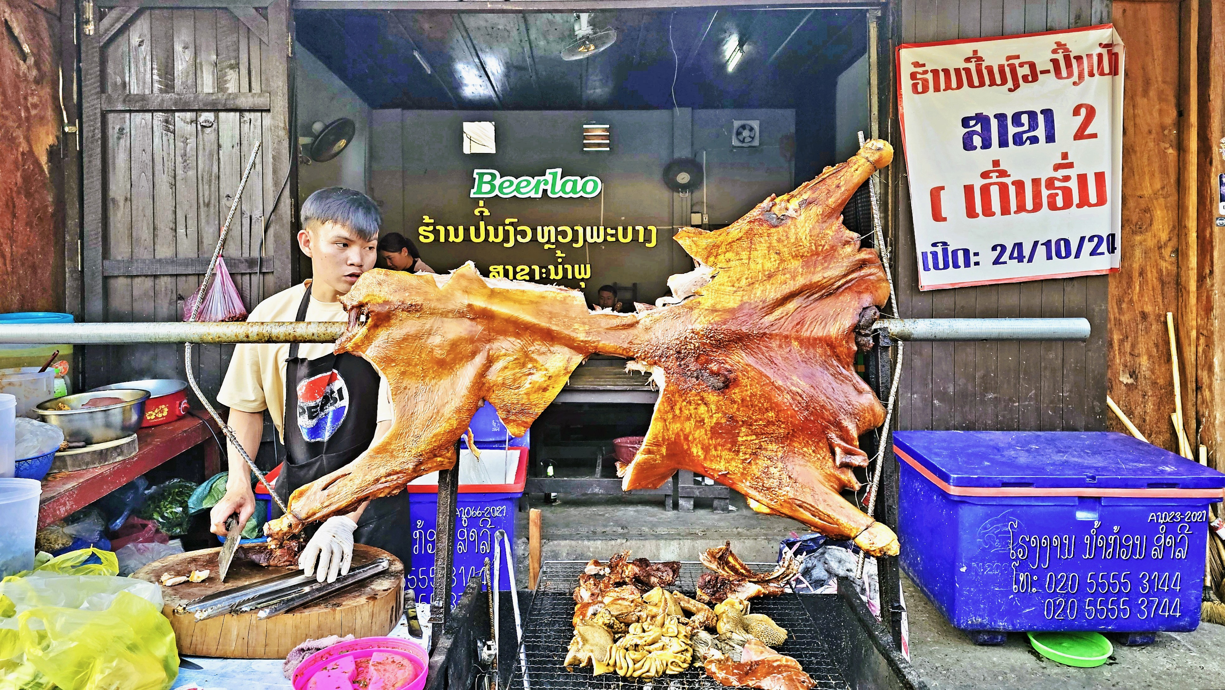 A vendor stands behind a stall with roasted meat, including pig and various dishes, in a local market setting.