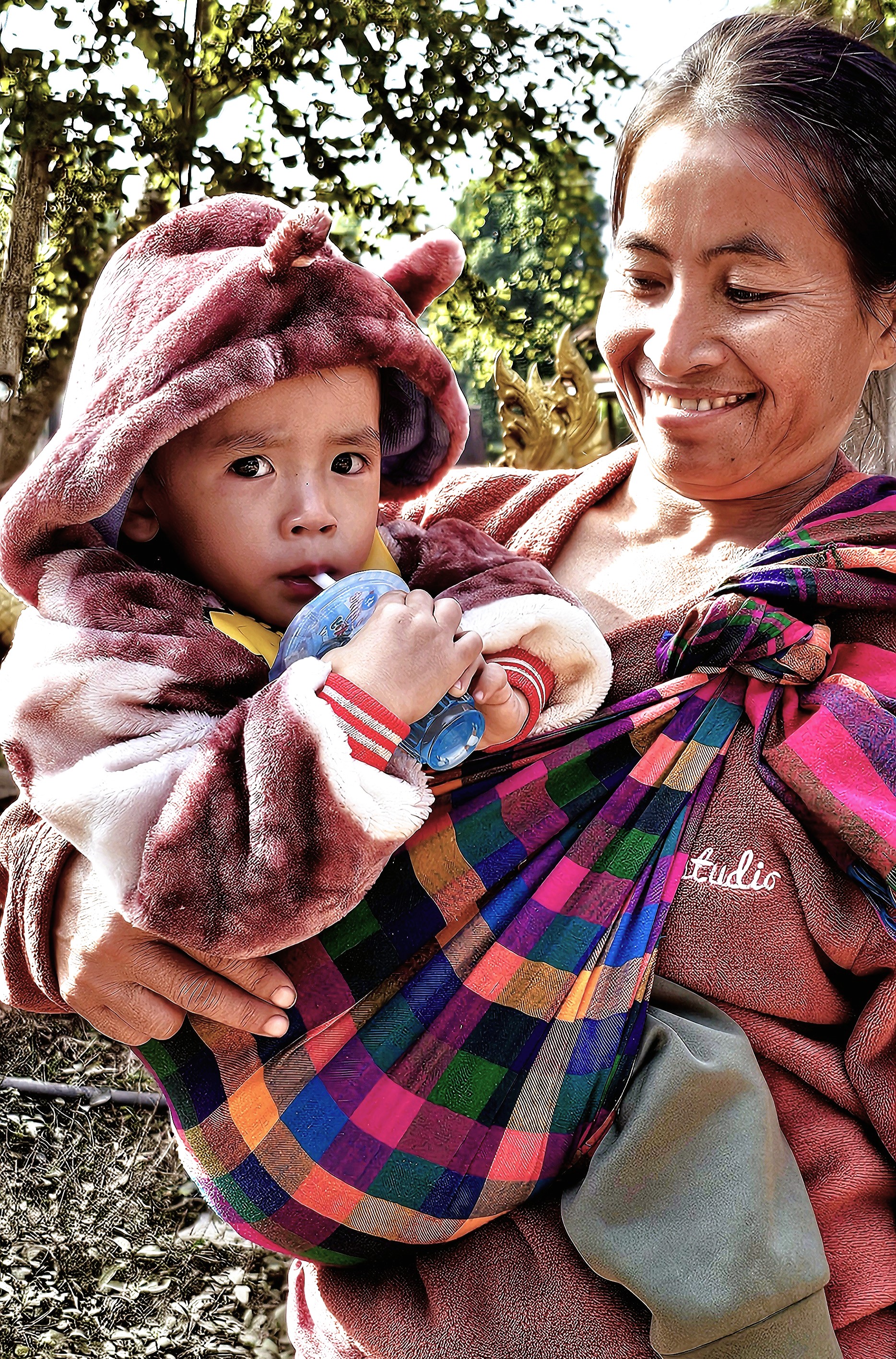A smiling woman holds a young child dressed in a patterned outfit, while the child clutches a plastic bottle, surrounded by nature.