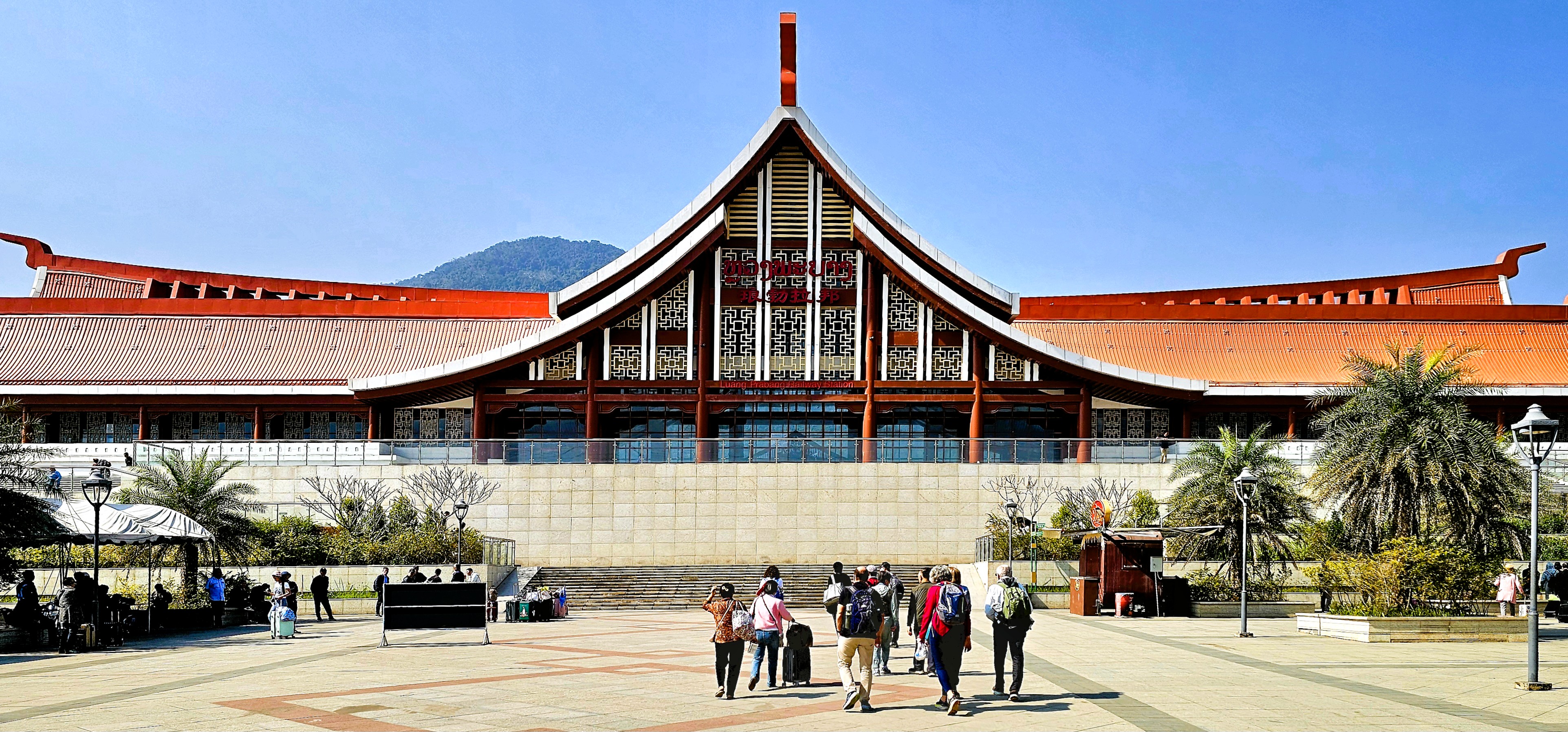 A group of travelers walking towards a modern building with a traditional Lao architectural style, featuring a curved roof and decorative elements, set against a clear blue sky.