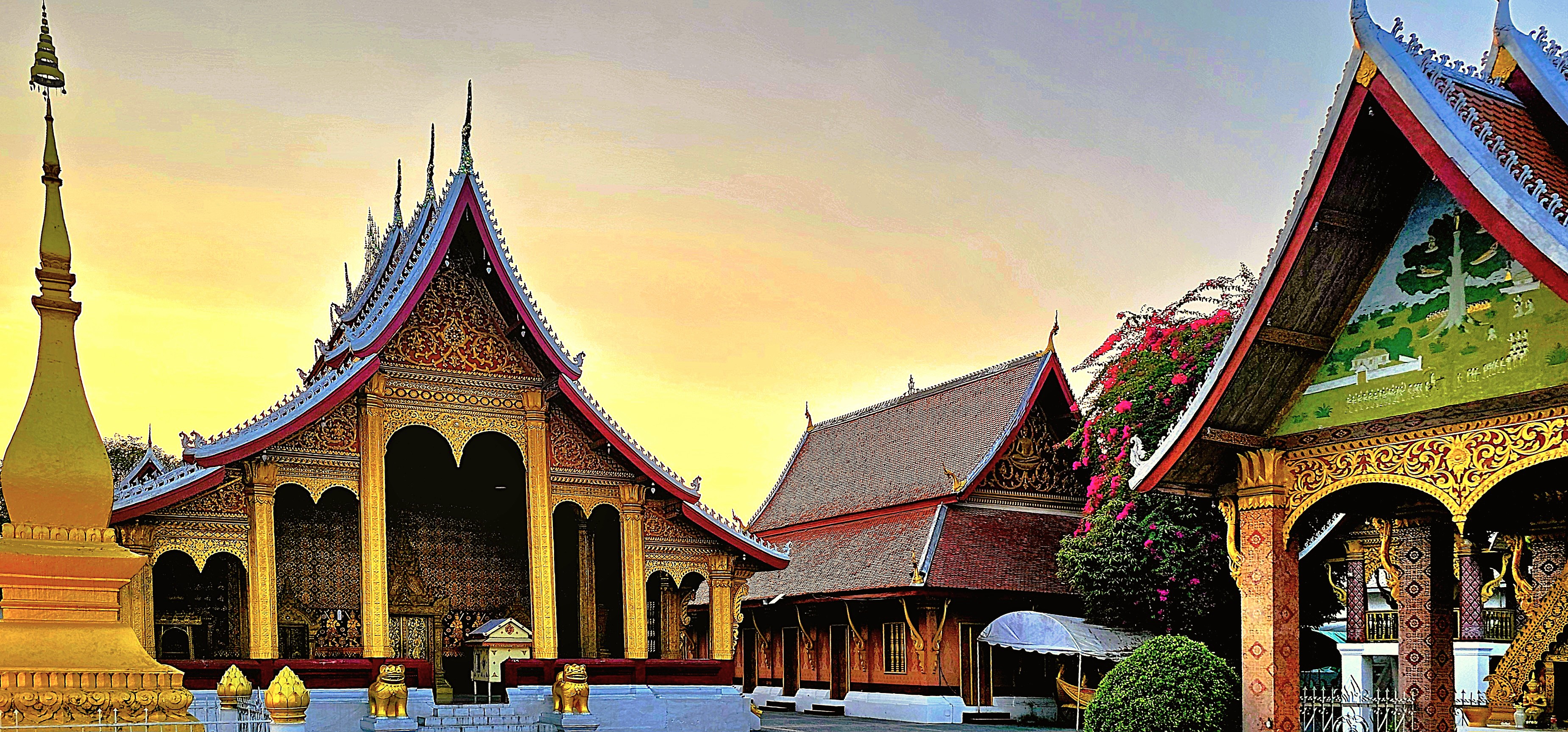 Sunset view of the ornate rooftops of traditional Lao temples, featuring intricate designs and vibrant colors, surrounded by greenery.