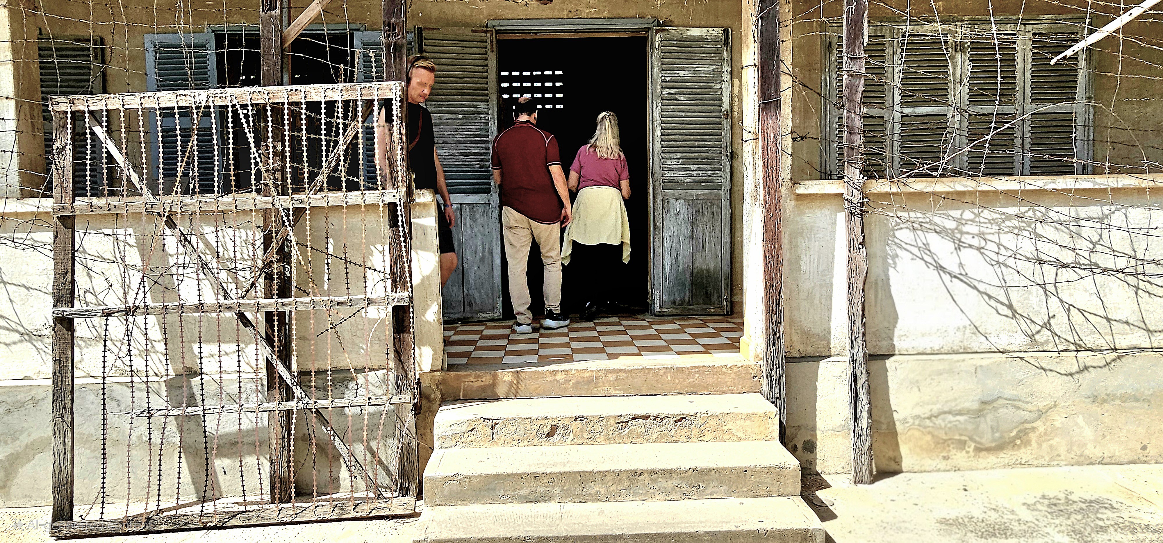 Visitors entering the Tuol Sleng Genocide Museum, formerly a school turned detention center, with barbed wire fencing visible in the foreground.