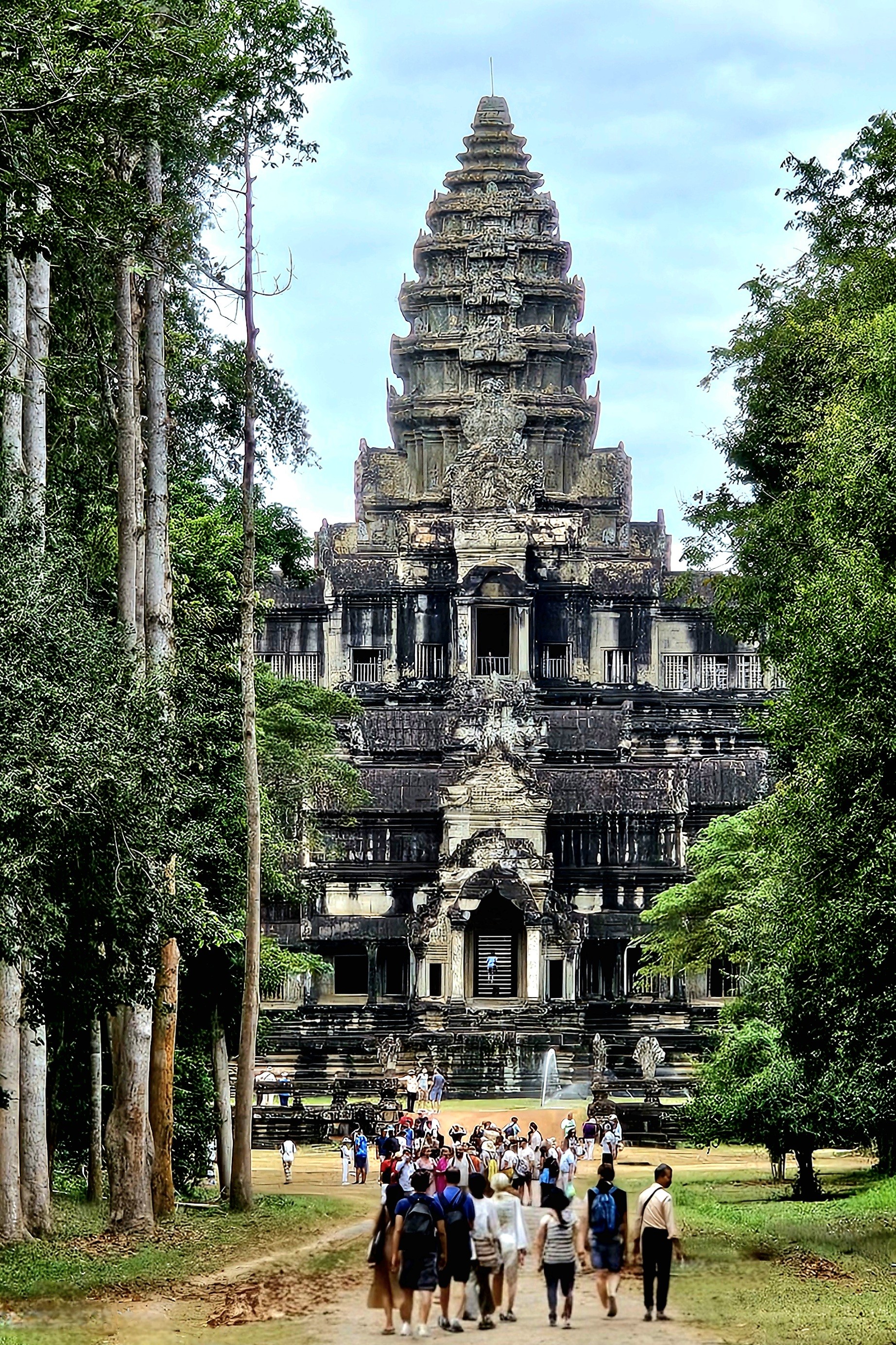 A view of Angkor Wat, a grand Khmer temple, with a crowd of visitors walking towards its entrance, surrounded by trees and greenery.