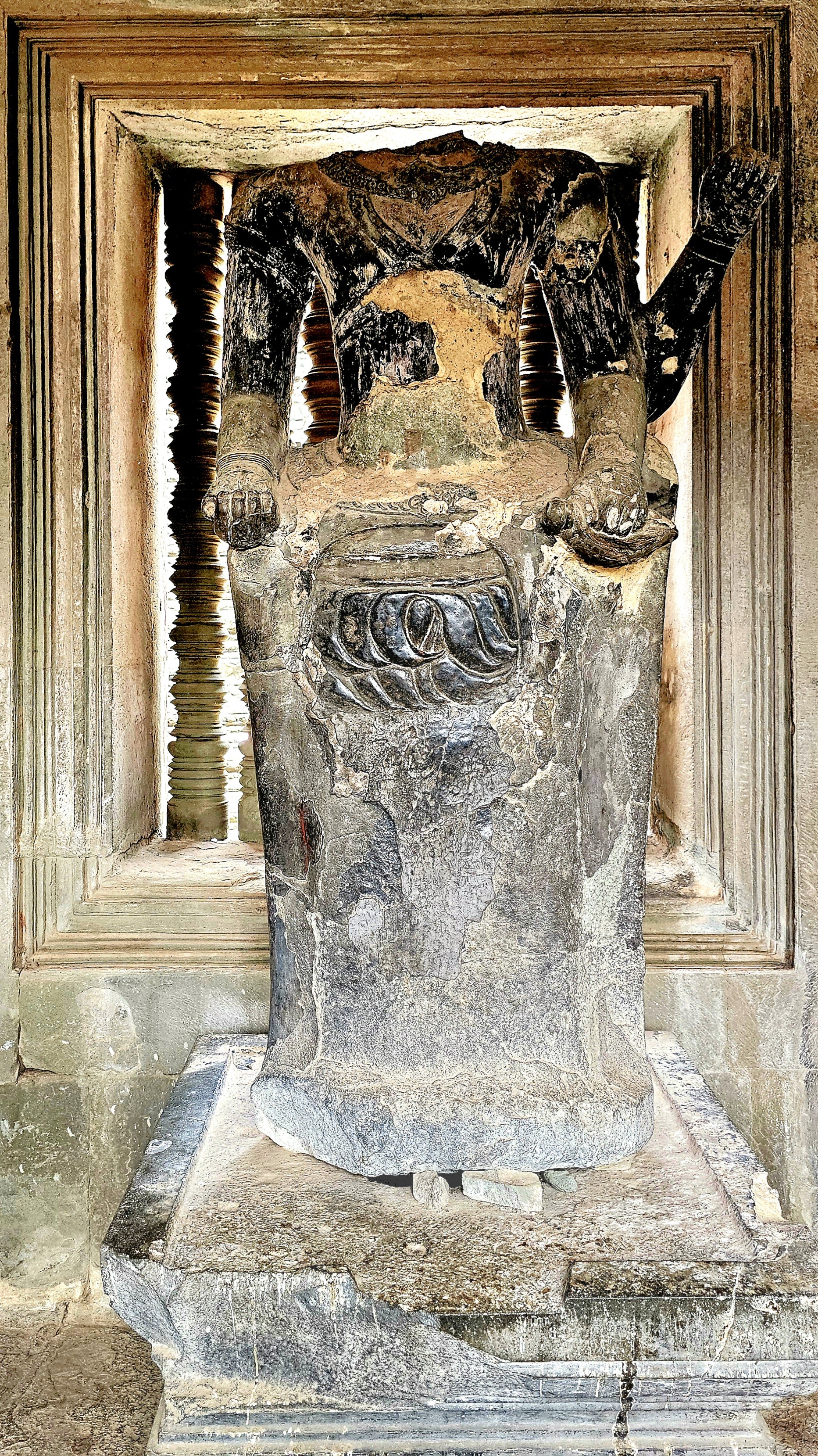 A partially eroded statue with missing head and arms, set against an ancient stone backdrop, showcasing intricate designs typical of Khmer art.
