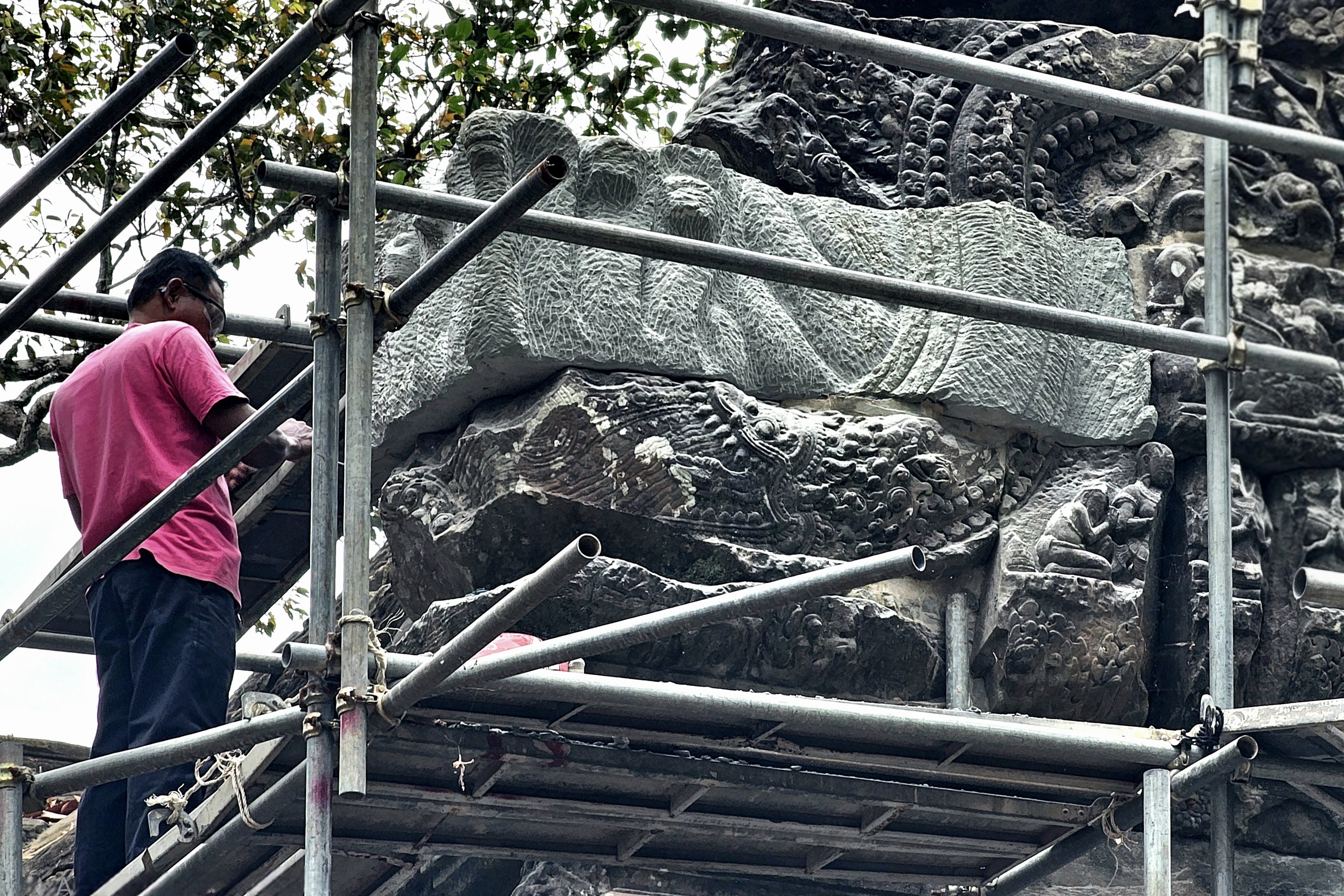 A worker is restoring intricate stone carvings on a temple in Siem Reap, Cambodia, using scaffolding for support.