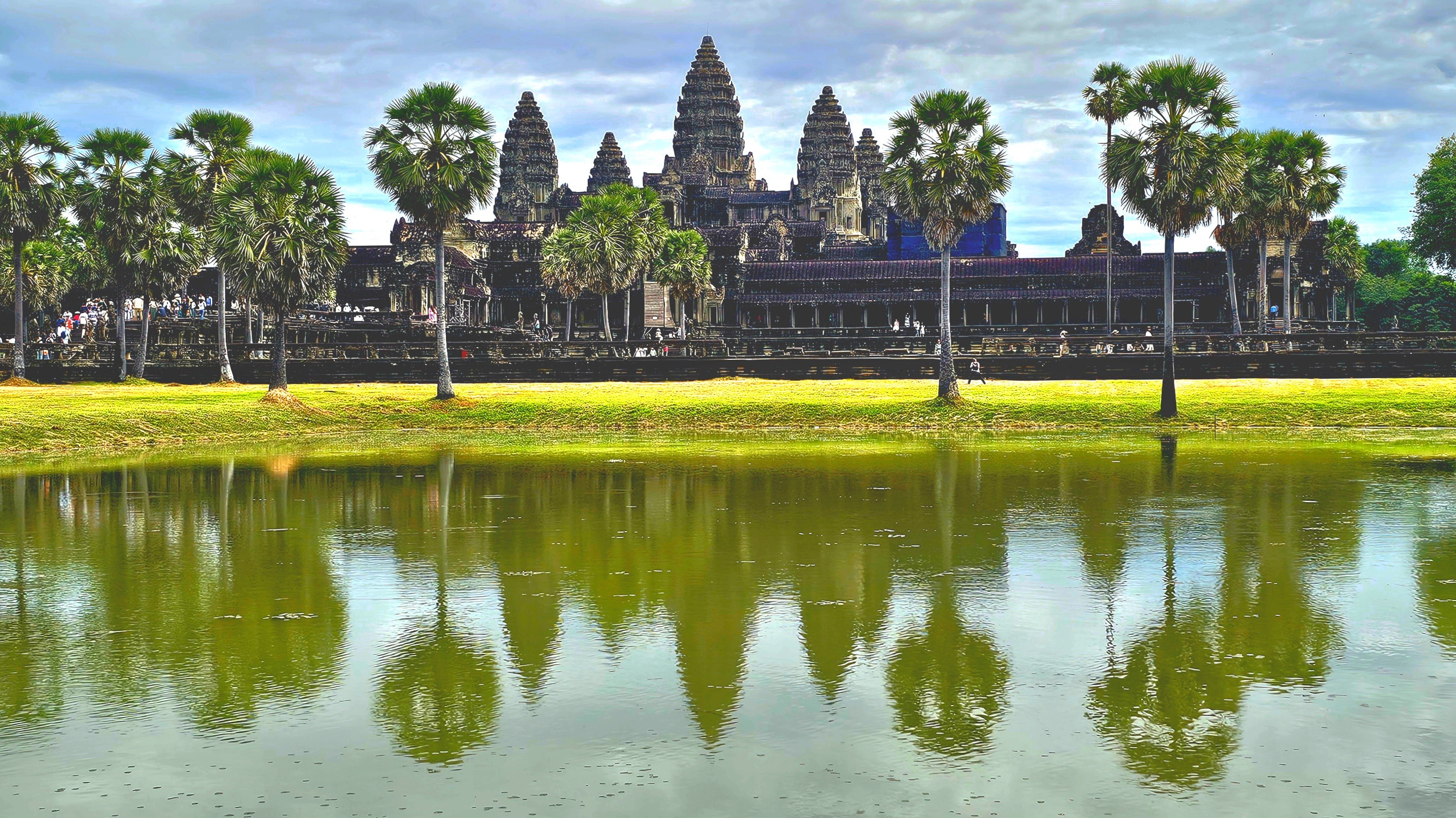 View of Angkor Wat temple, with lush greenery and palm trees reflected in the water of the surrounding moat.