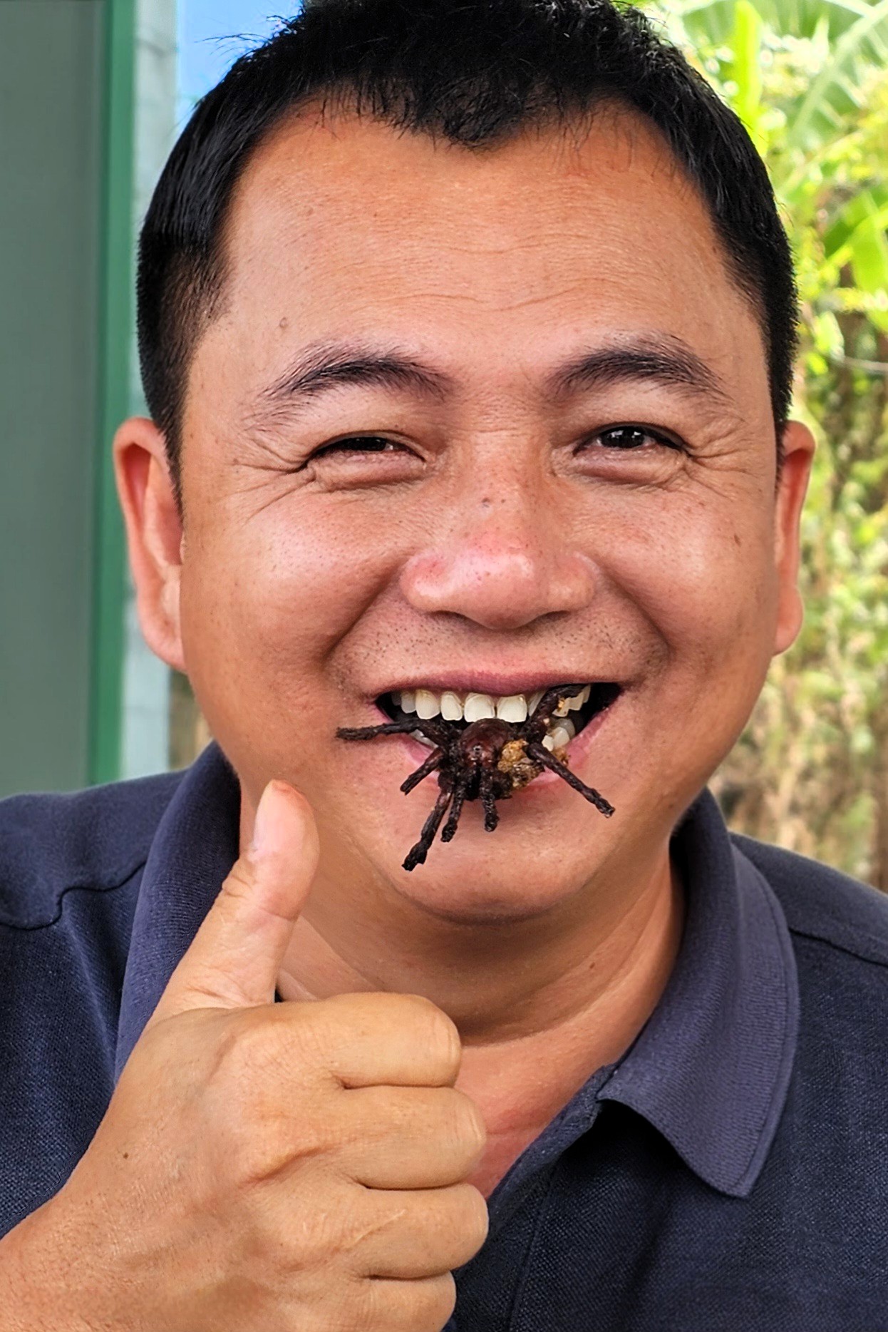 A man smiling and giving a thumbs-up while holding a fried tarantula in his mouth, showcasing local cuisine in Cambodia.