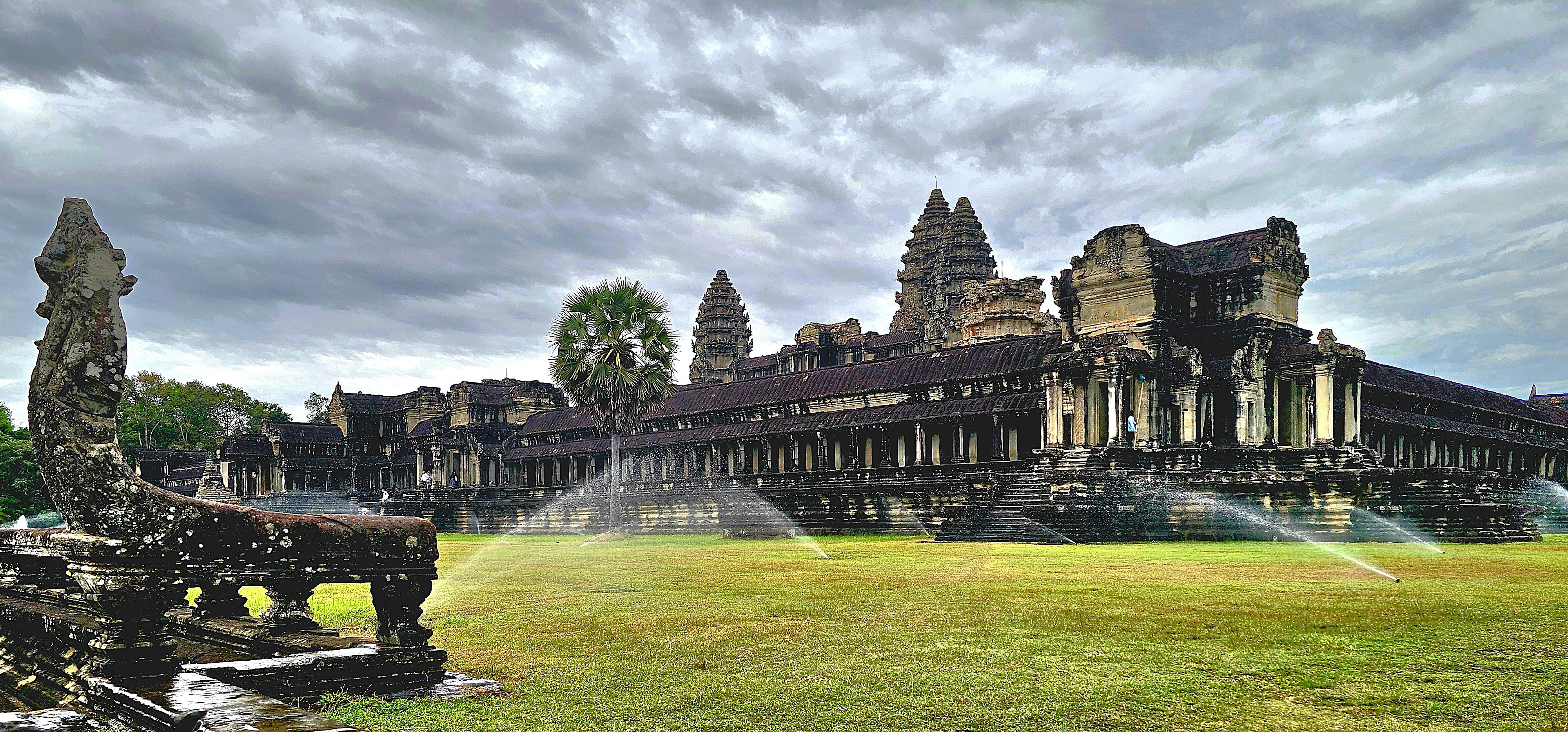 A view of Angkor Wat under cloudy skies, featuring the temple's distinct architecture with its multiple spires and a nearby stone naga statue in the foreground.