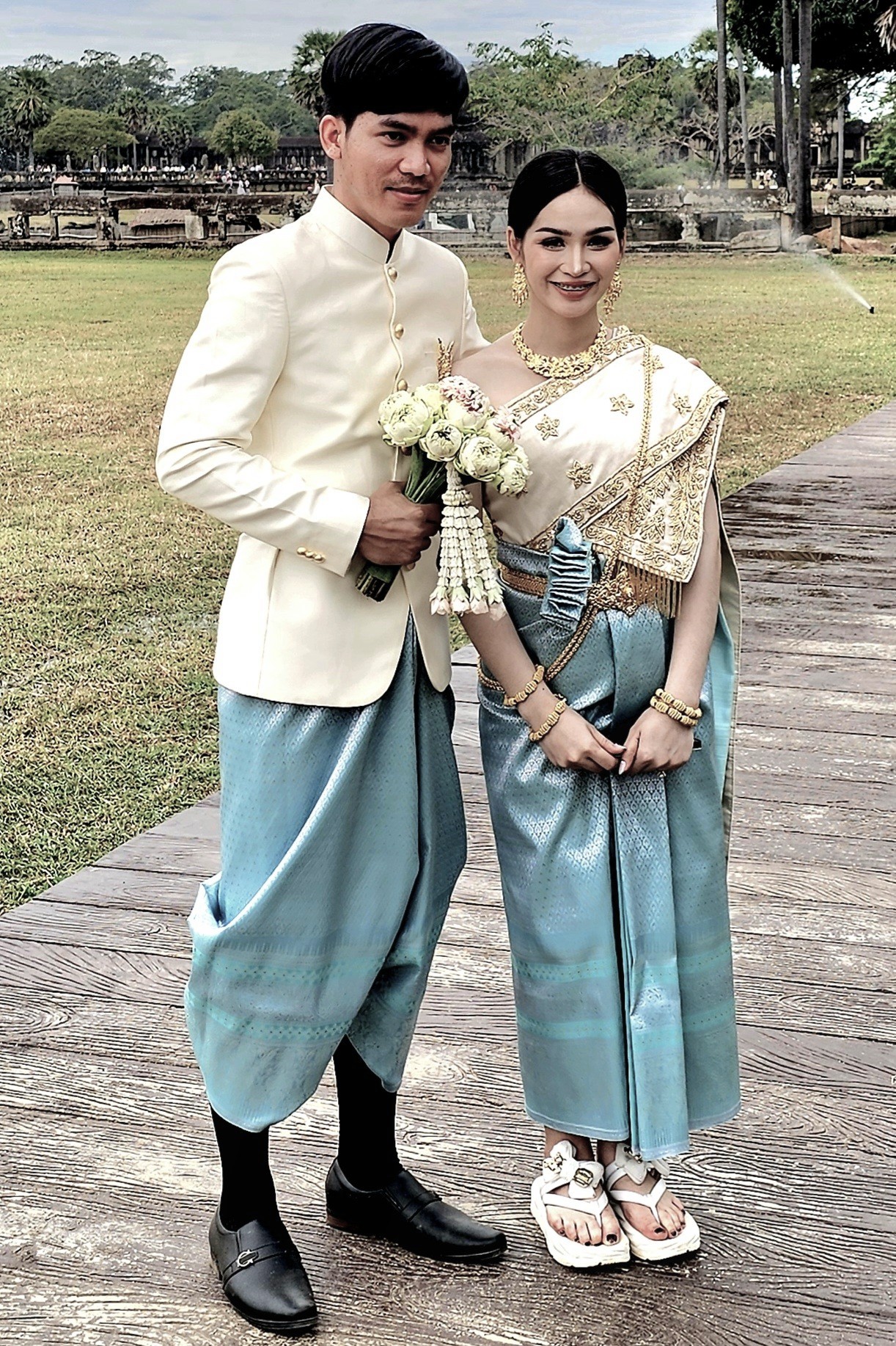 A man and a woman in traditional Cambodian attire, posing together with a bouquet of flowers, against a scenic backdrop that hints at the cultural significance of their location.