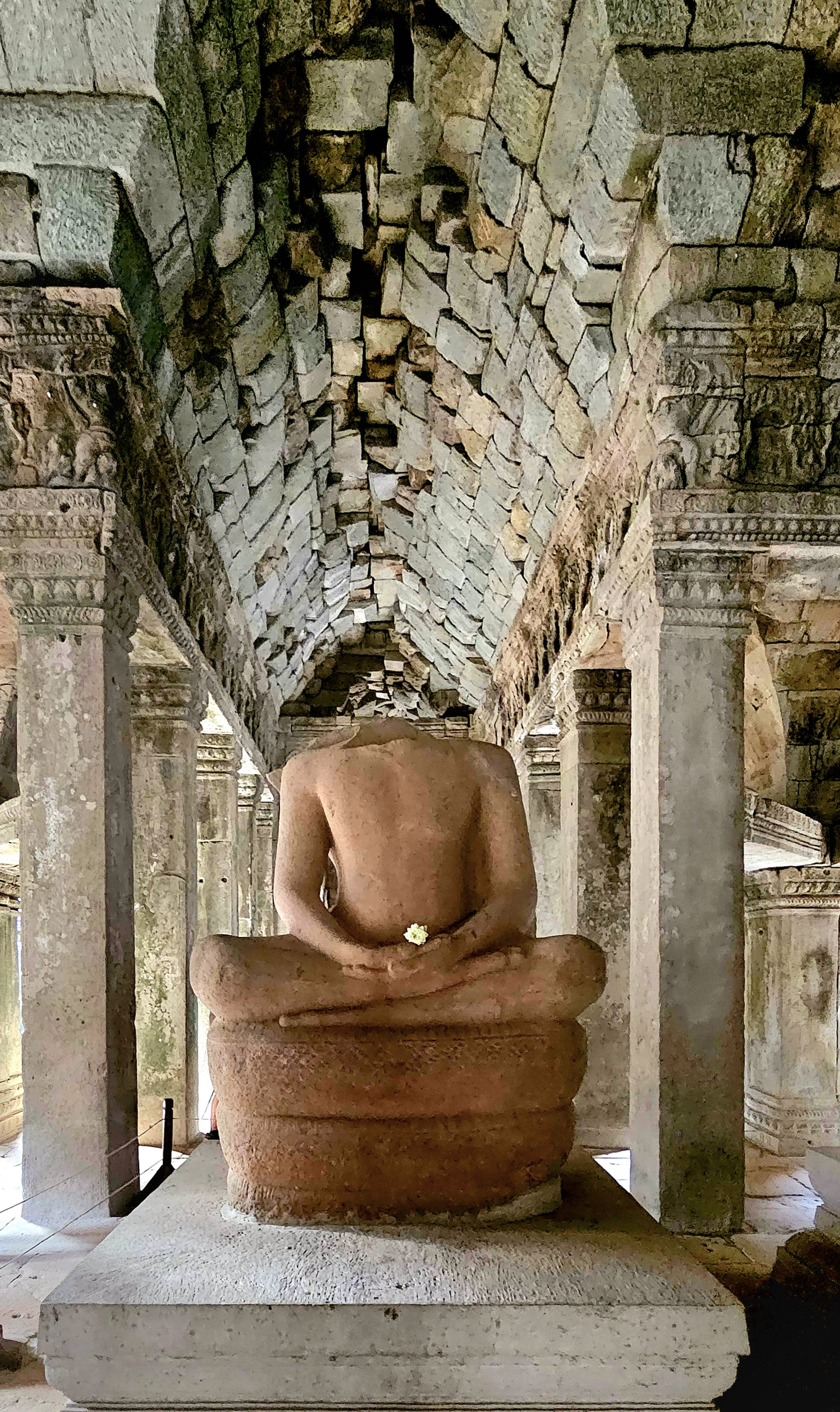 A stone statue of a seated Buddha, viewed from the back, inside a temple with intricately decorated columns and a tiled ceiling.