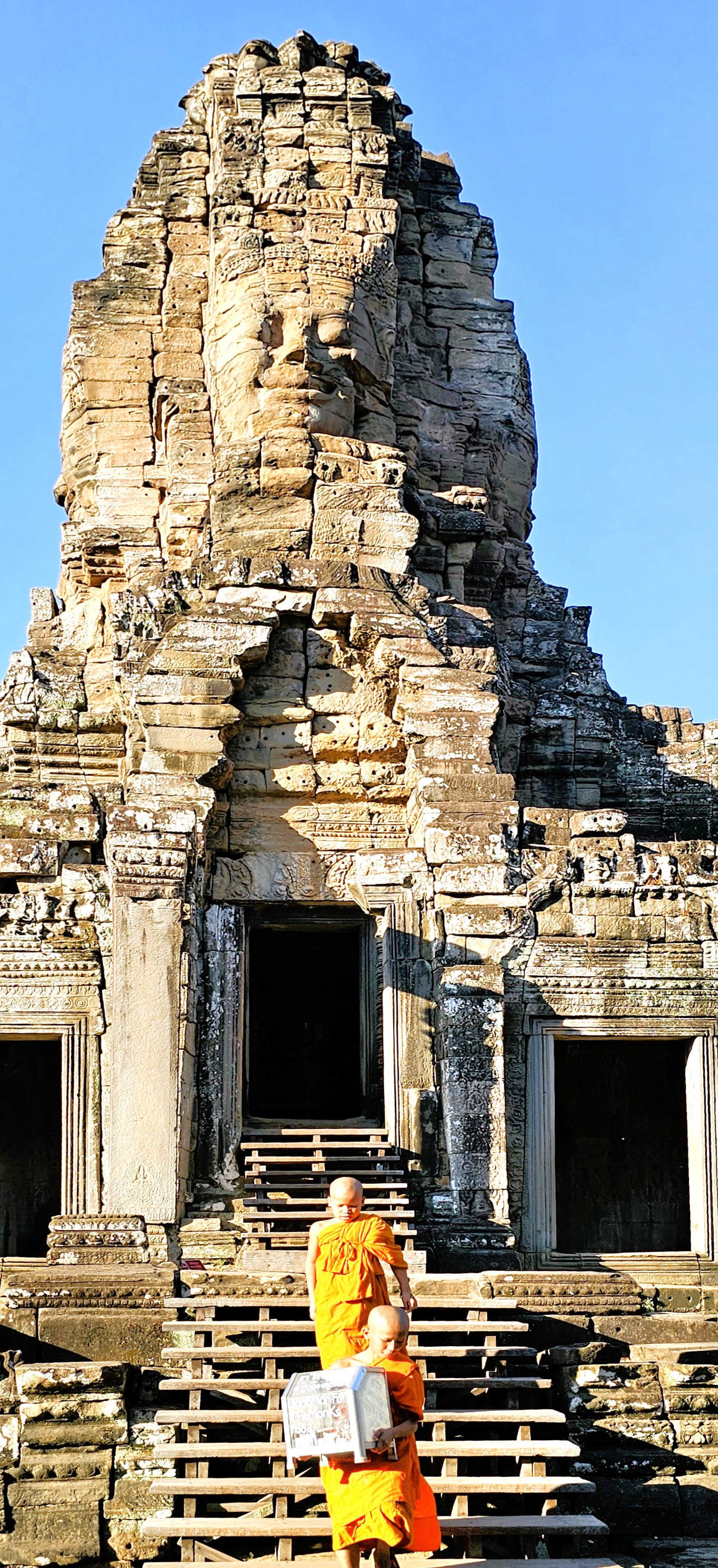 Two young monks in orange robes descending the stairs of Bayon Temple, featuring a towering stone face in the background on a clear blue sky.
