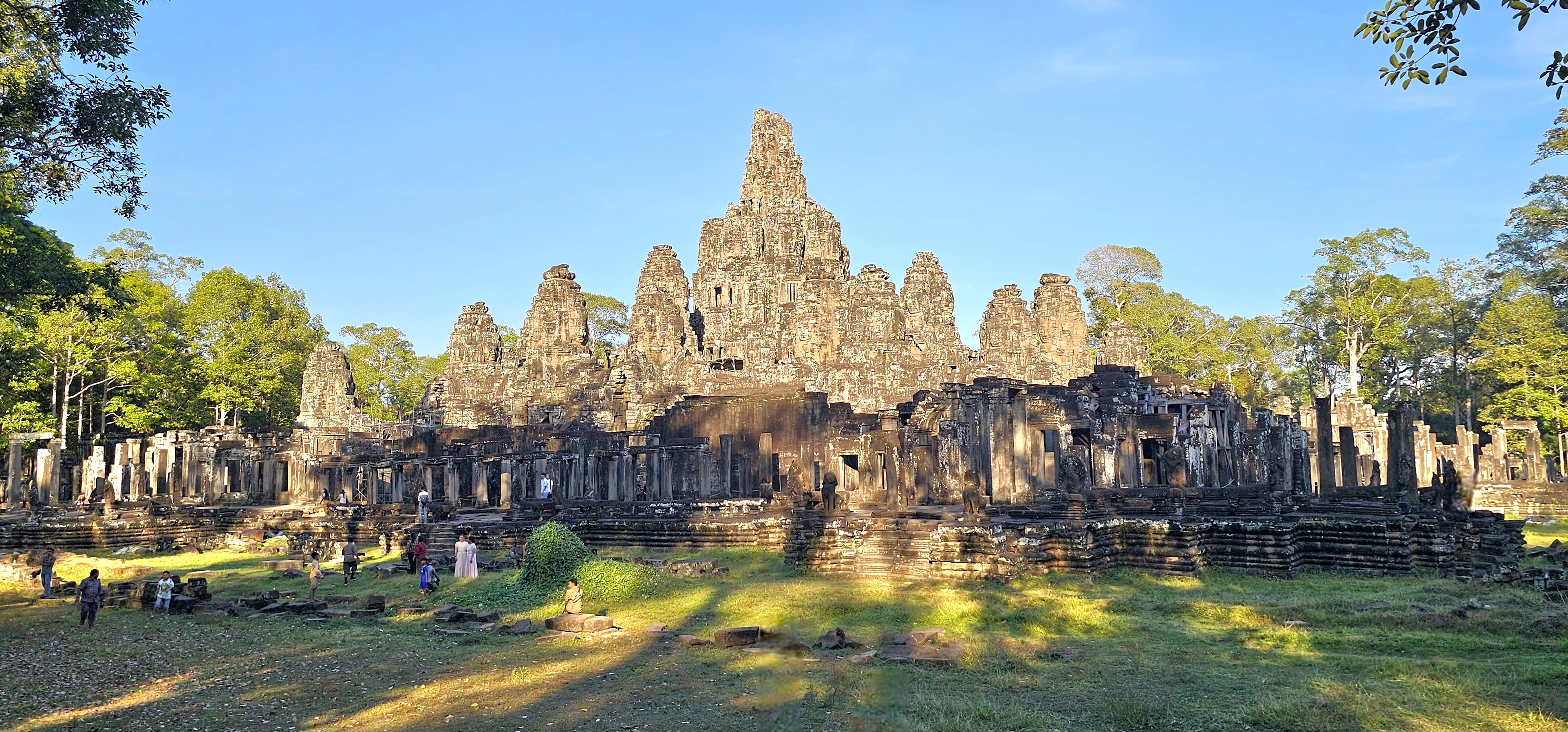 A panoramic view of Bayon Temple in Angkor Thom, showcasing its distinctive towers amidst lush greenery and a clear blue sky, with visitors exploring the ancient structure.