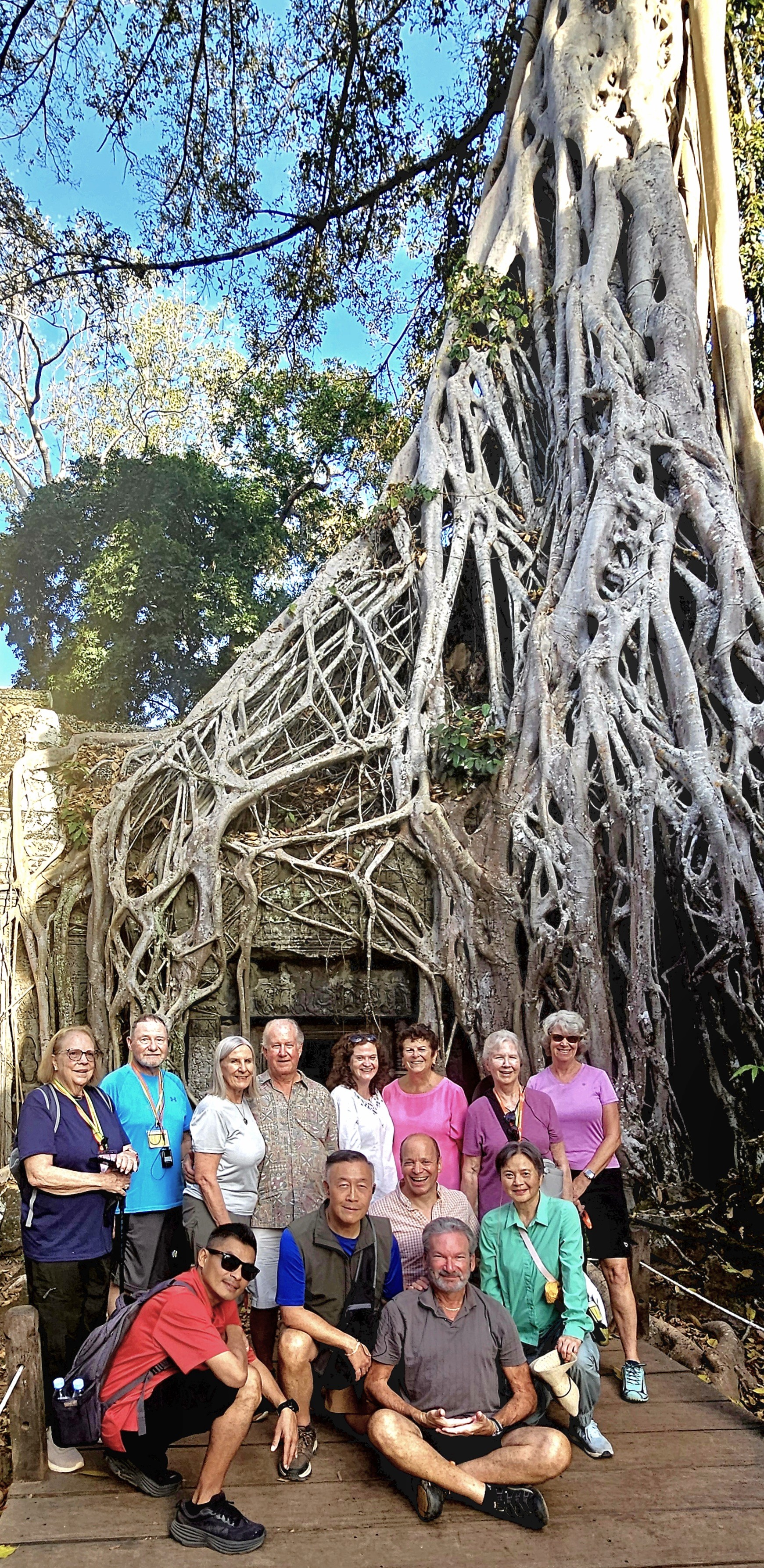 A group of tourists poses in front of ancient stone ruins enveloped by a massive tree's root system at Ta Prohm temple in Cambodia.
