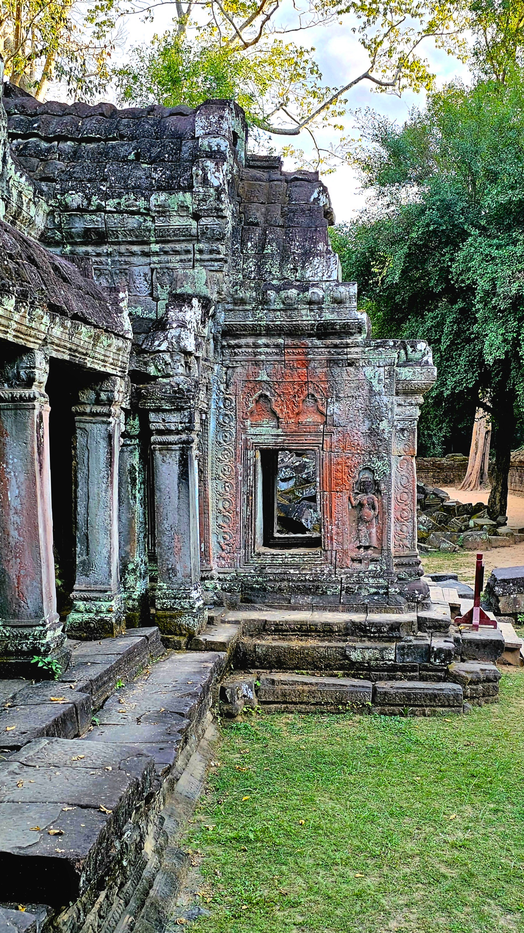 A partially restored structure of the Ta Prohm temple in Cambodia, featuring intricate carvings and a doorway framed by stone columns, with greenery in the background.