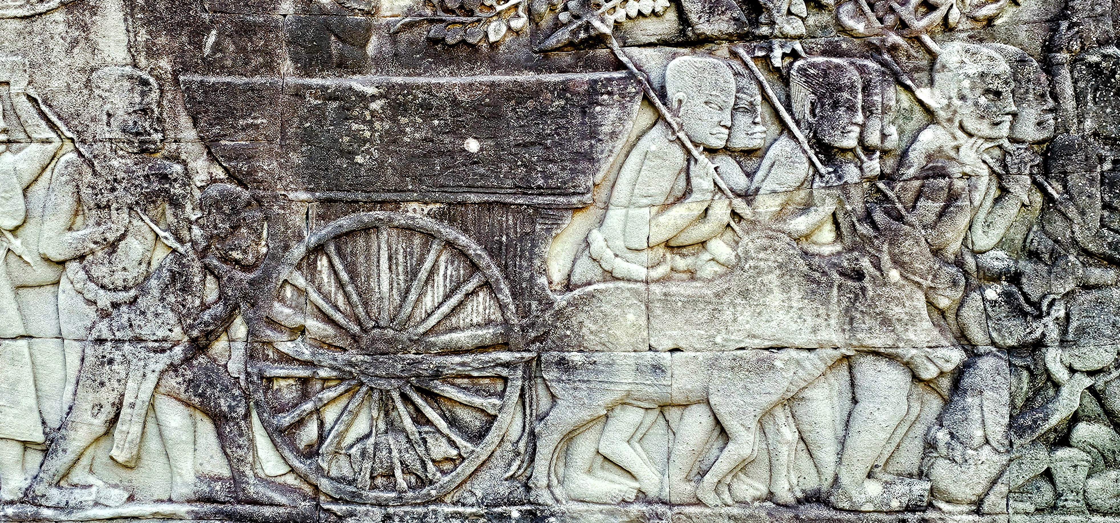 Detailed relief sculpture depicting historical battle scenes and everyday life, featuring figures with weapons, a cart, and animals, carved into stone at Bayon Temple.