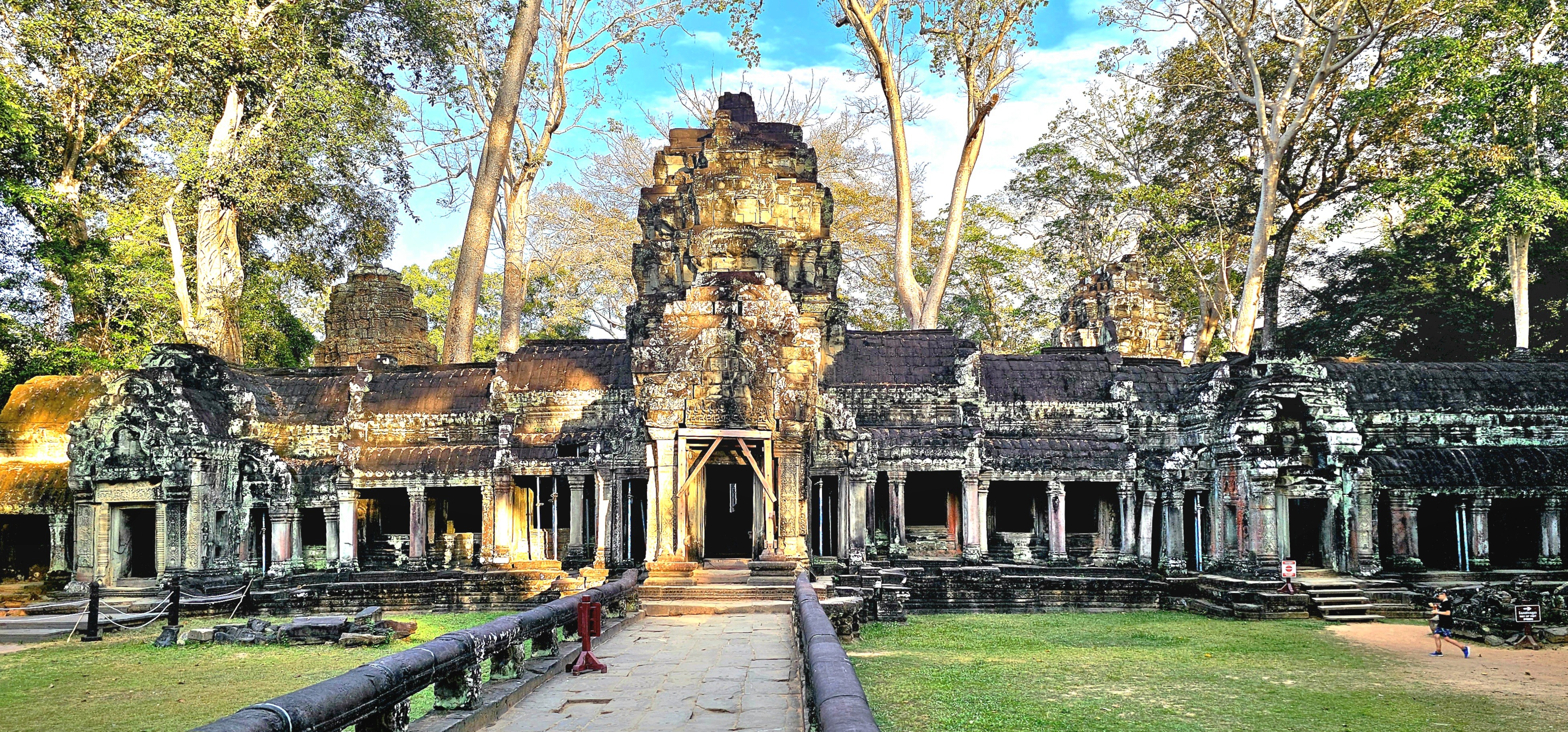 A view of the Ta Prohm temple in Angkor Archaeological Park, showcasing ancient stone structures intertwined with large tree roots and surrounded by lush greenery.