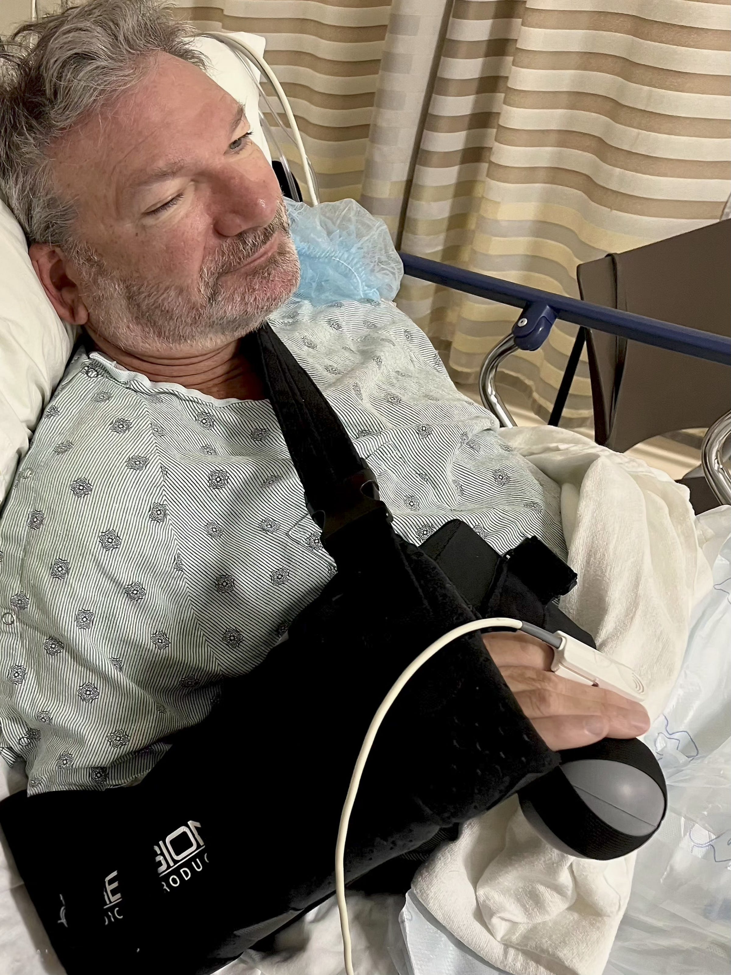 A man in a hospital bed with a surgical gown and a black arm sling, looking relaxed with a slight smile. Medical equipment is visible in the background.