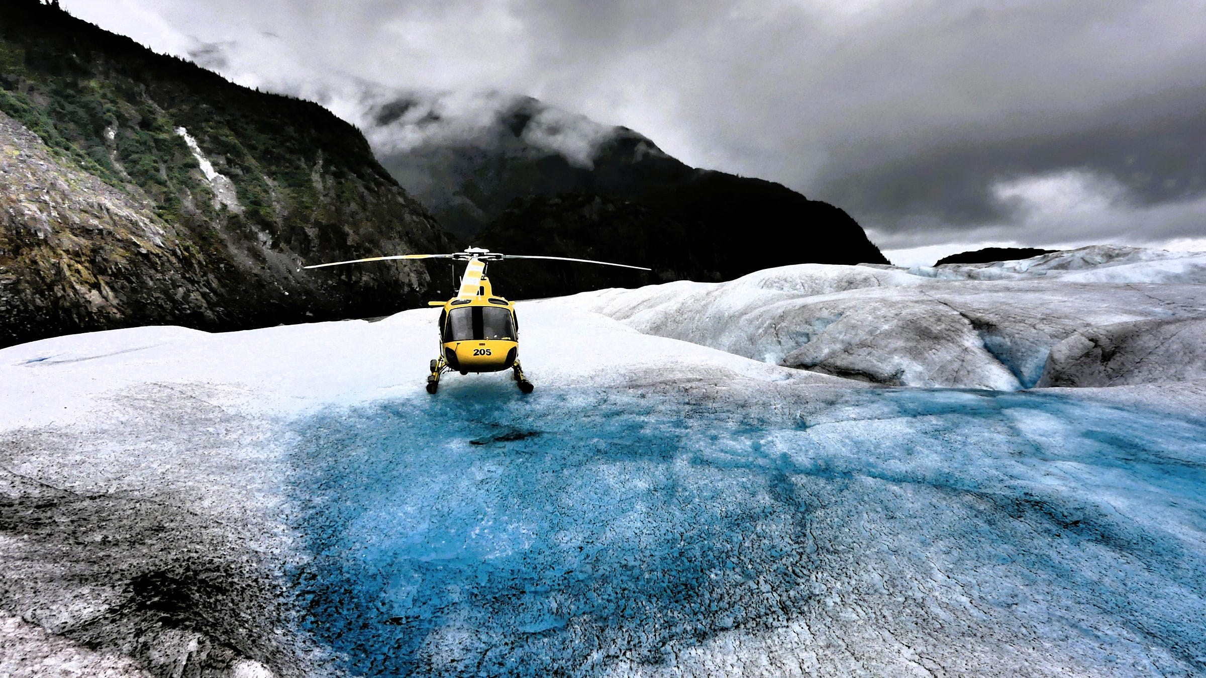 A yellow helicopter on a glacier, surrounded by dramatic mountains under a cloudy sky.