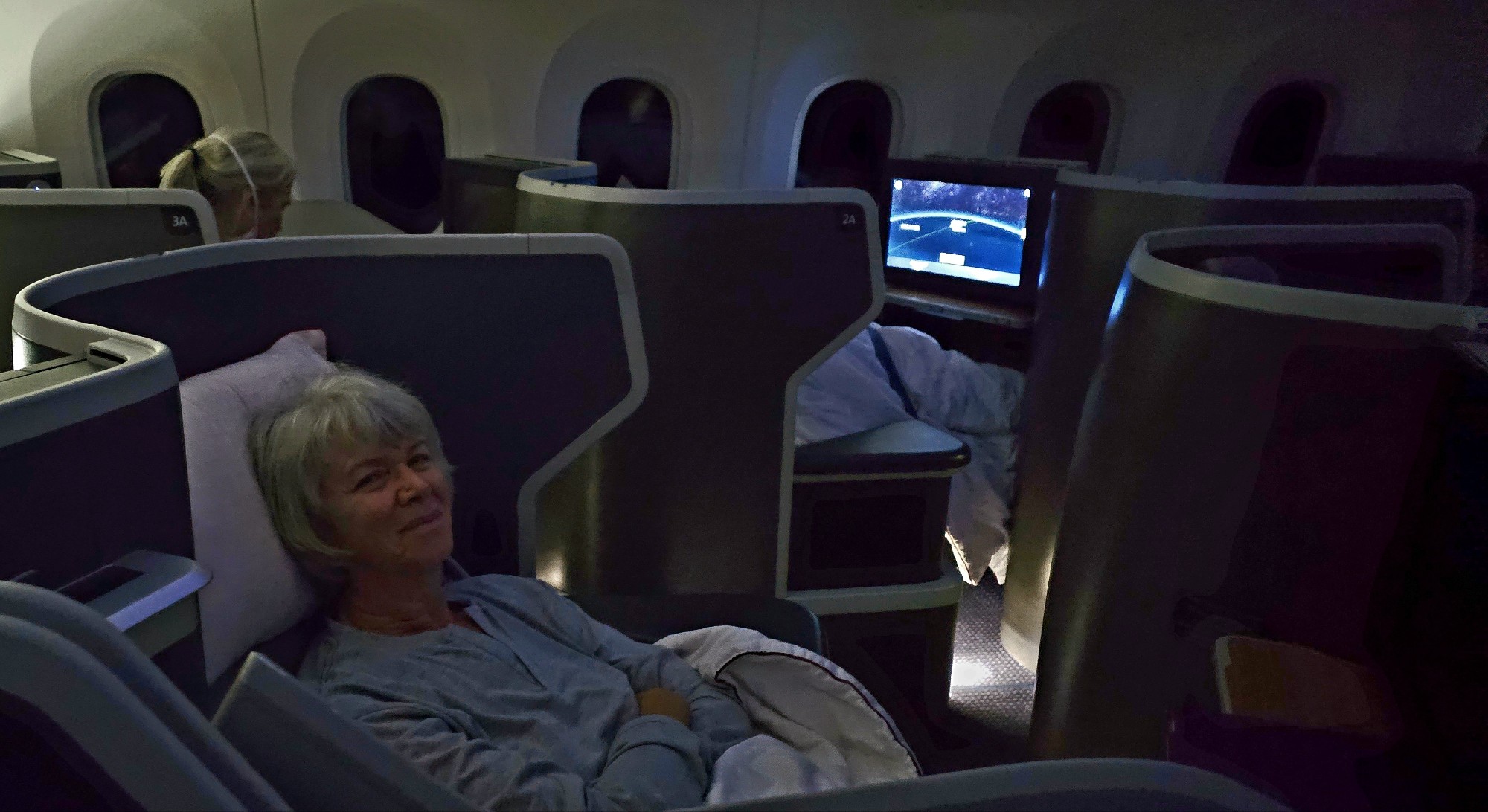 A passenger with gray hair sits comfortably in a business class airplane seat during a night flight, with a personal entertainment screen showing a starry sky.