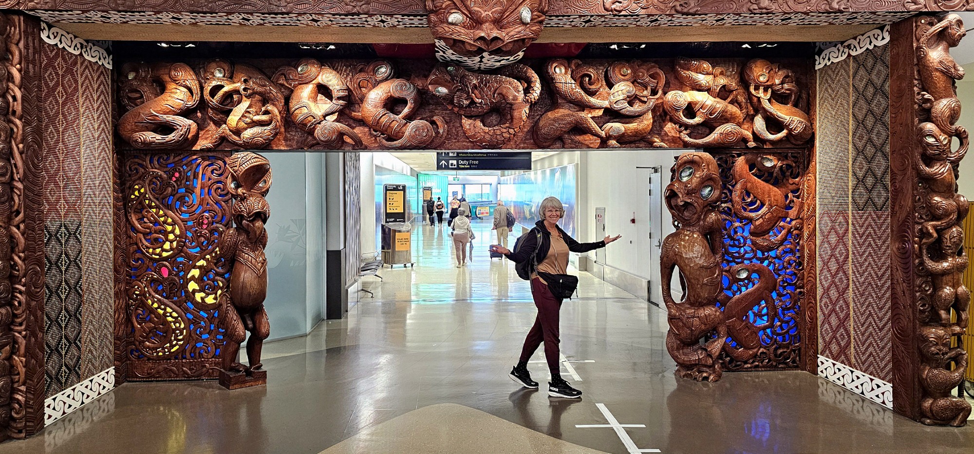 A person stands in front of a large, intricately carved wooden archway adorned with various figures and designs at an airport. The background shows people walking down a terminal.