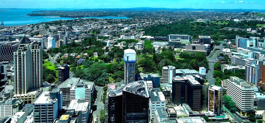 Aerial view of a cityscape featuring tall buildings, green parks, and a coastline with blue waters in the background.