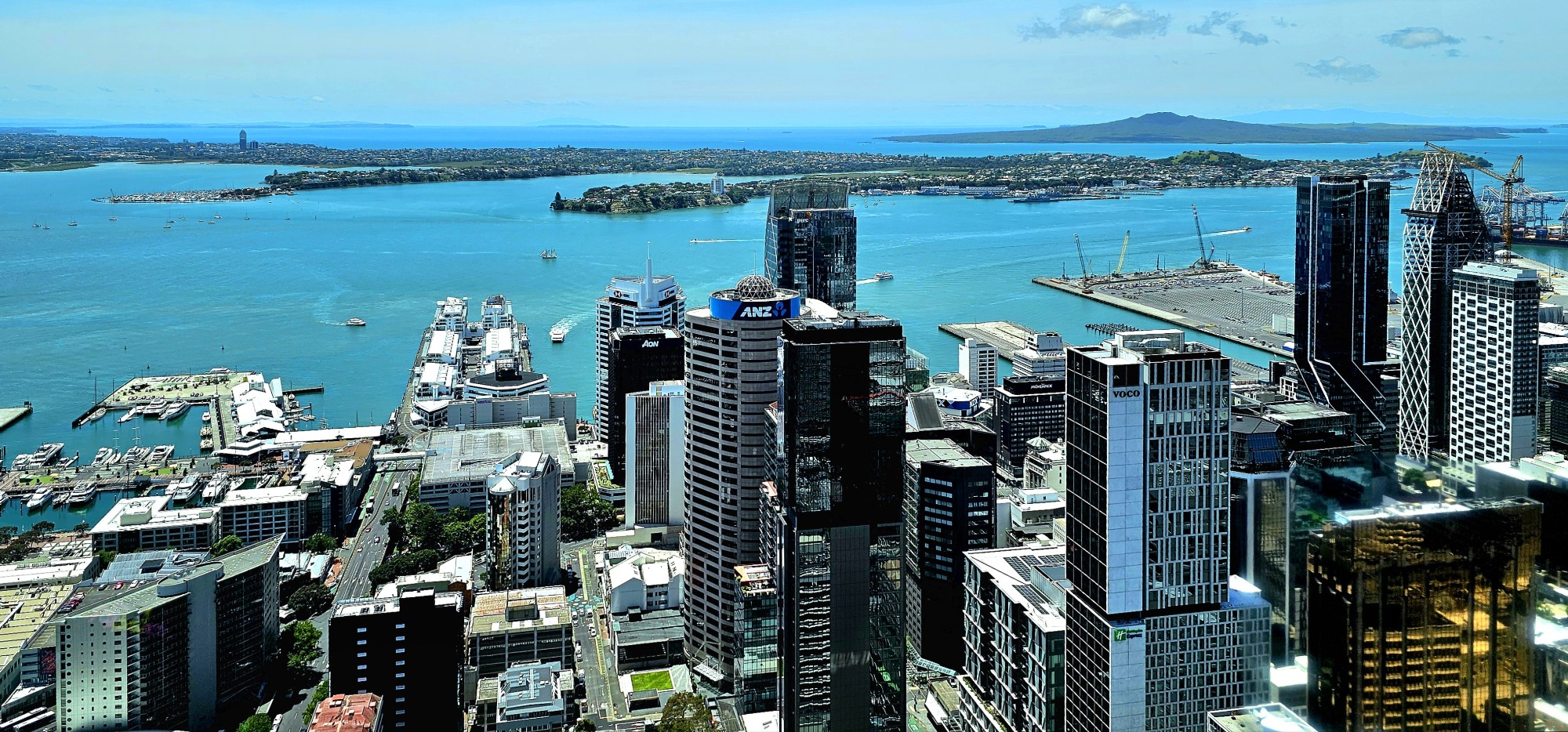 A panoramic view of a city skyline with modern skyscrapers beside a turquoise harbor, featuring boats and distant islands under a clear blue sky.