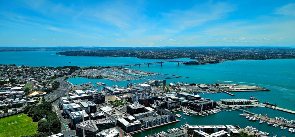 Aerial view of a coastal city featuring marinas, buildings, and a bridge over turquoise waters on a sunny day.