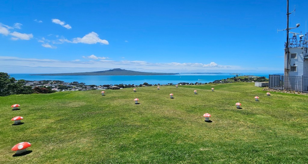 A scenic view of a green grassy area dotted with red and white mushrooms, overlooking a blue ocean and a distant island under a clear blue sky with a few clouds.