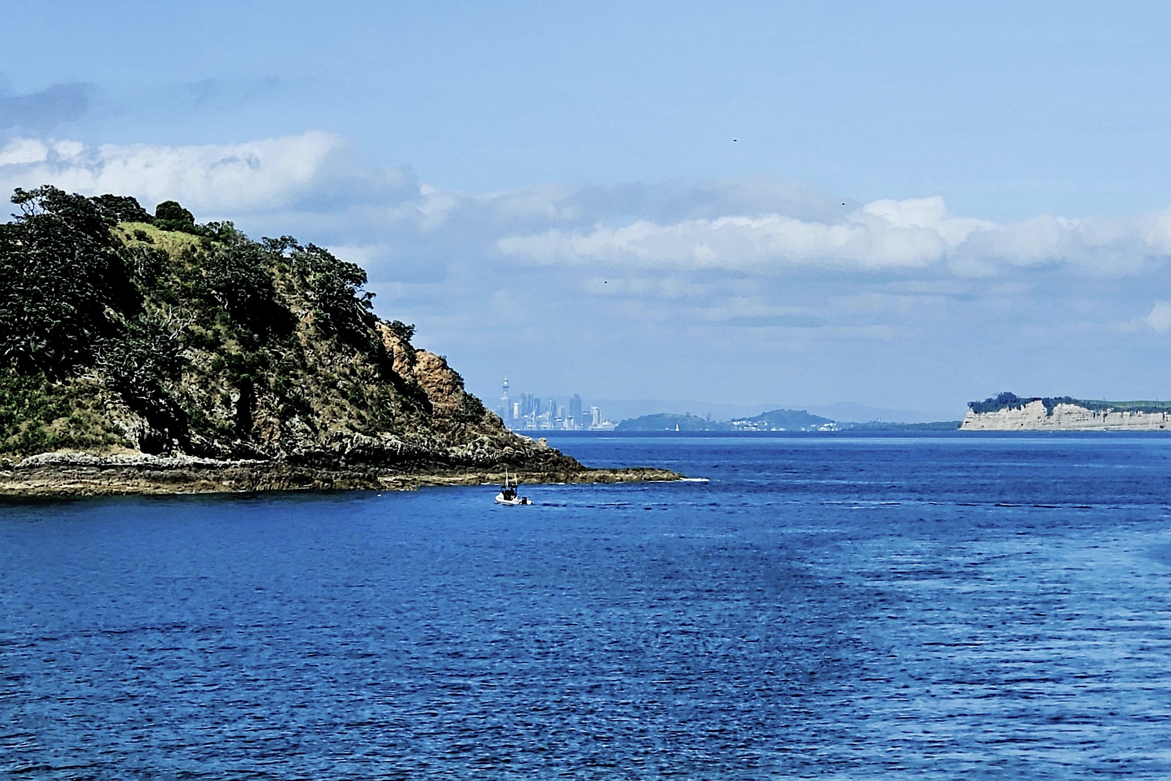 A scenic view of a coastal landscape featuring a rocky island with lush greenery, a small boat sailing in the calm blue waters, and a distant city skyline under a partly cloudy sky.