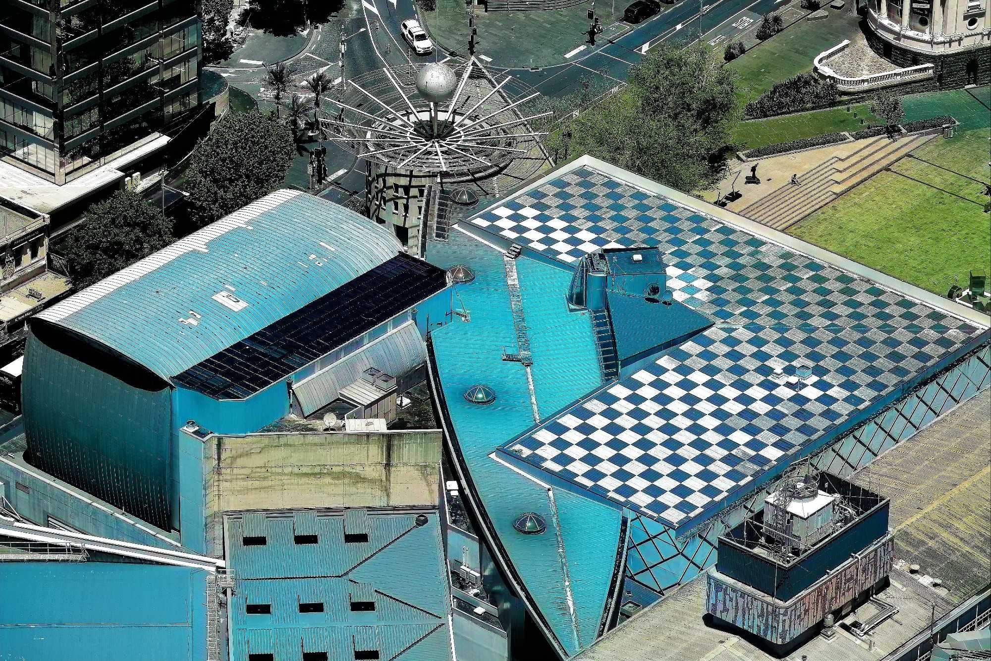Aerial view of a modern architectural complex featuring buildings with distinctive blue roofs, one with a checkered pattern and another with a curved design, surrounded by greenery and urban streets.
