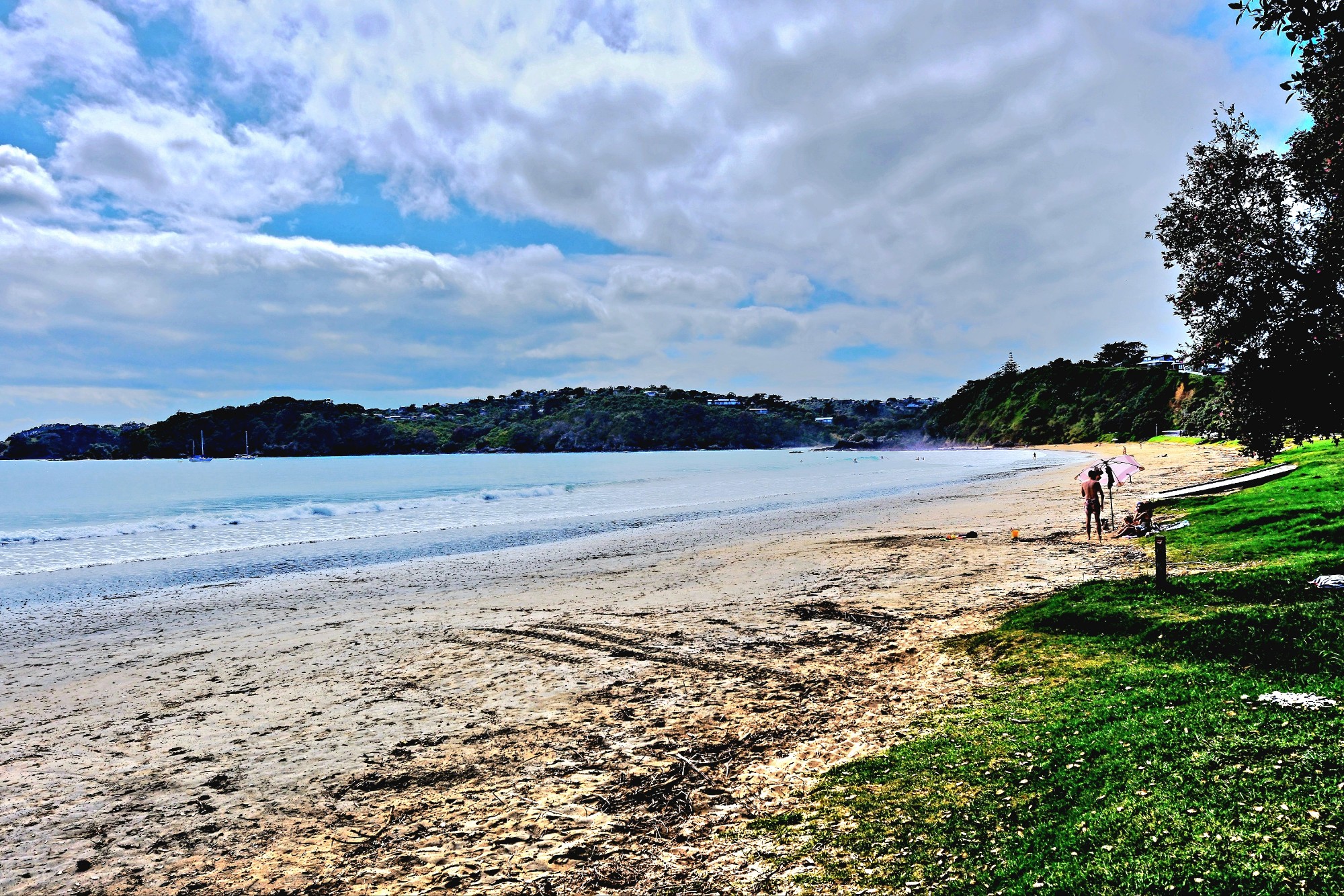 A panoramic view of a peaceful beach with soft sand, gentle waves, and green hills in the background. Two people are walking along the shore, and several boats are anchored in the water.