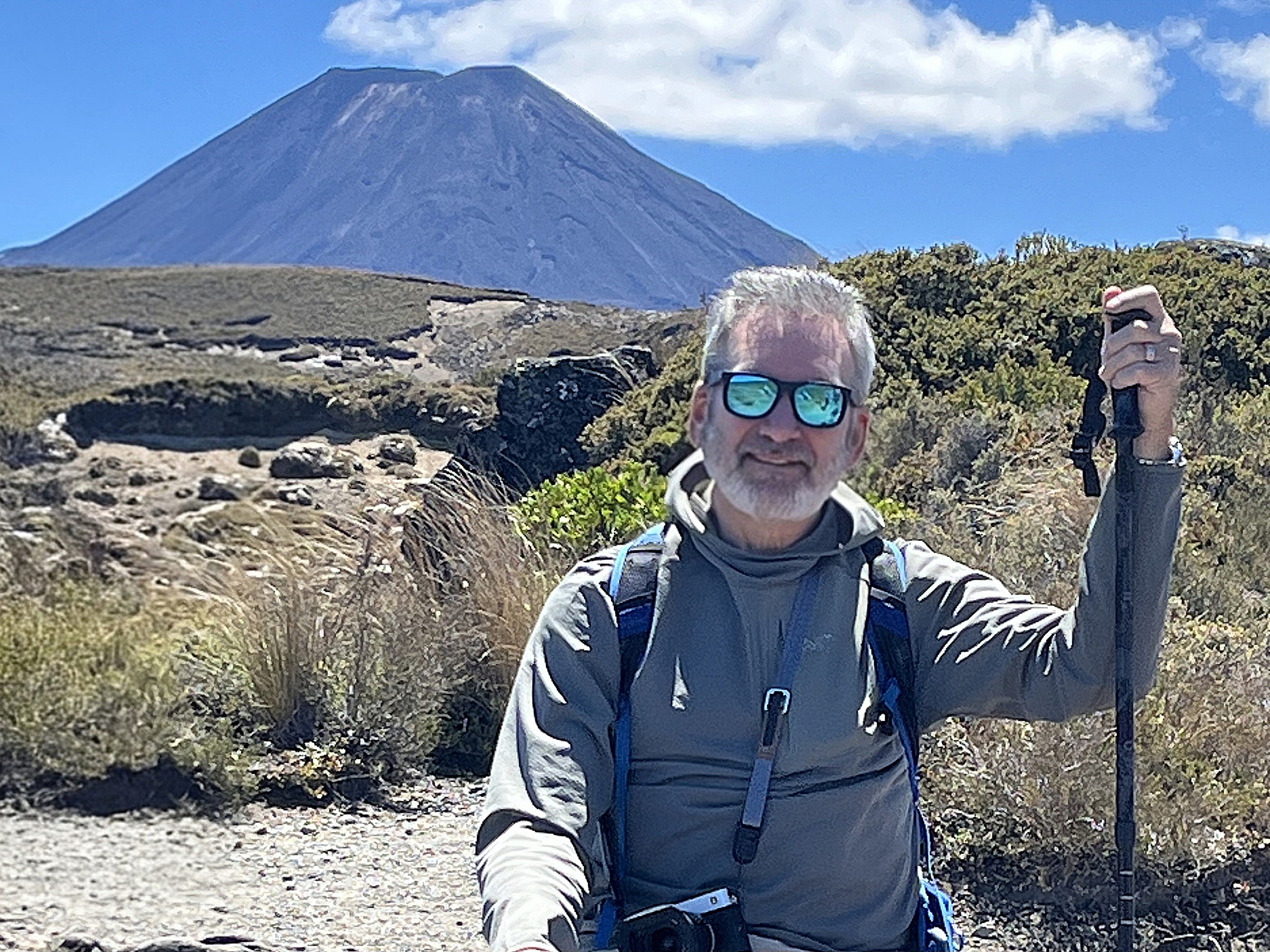 A smiling man wearing sunglasses and a gray hoodie sits on a hiking trail, holding a trekking pole, with a mountain in the background.