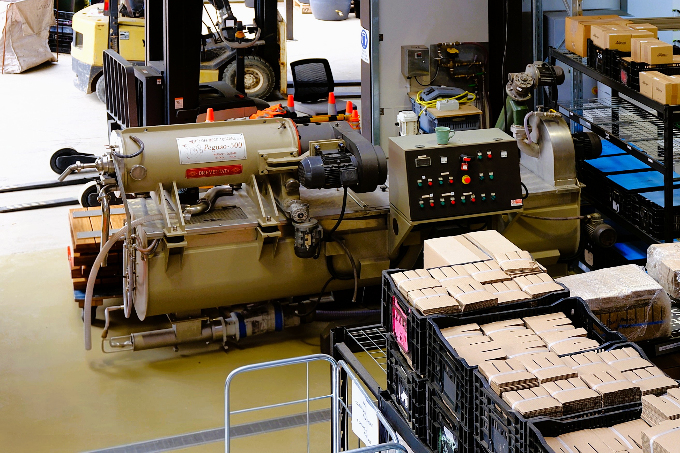 A production area featuring a large machine labeled 'Pegaso-500' with various control buttons, beside stacks of cardboard boxes stored in black crates.