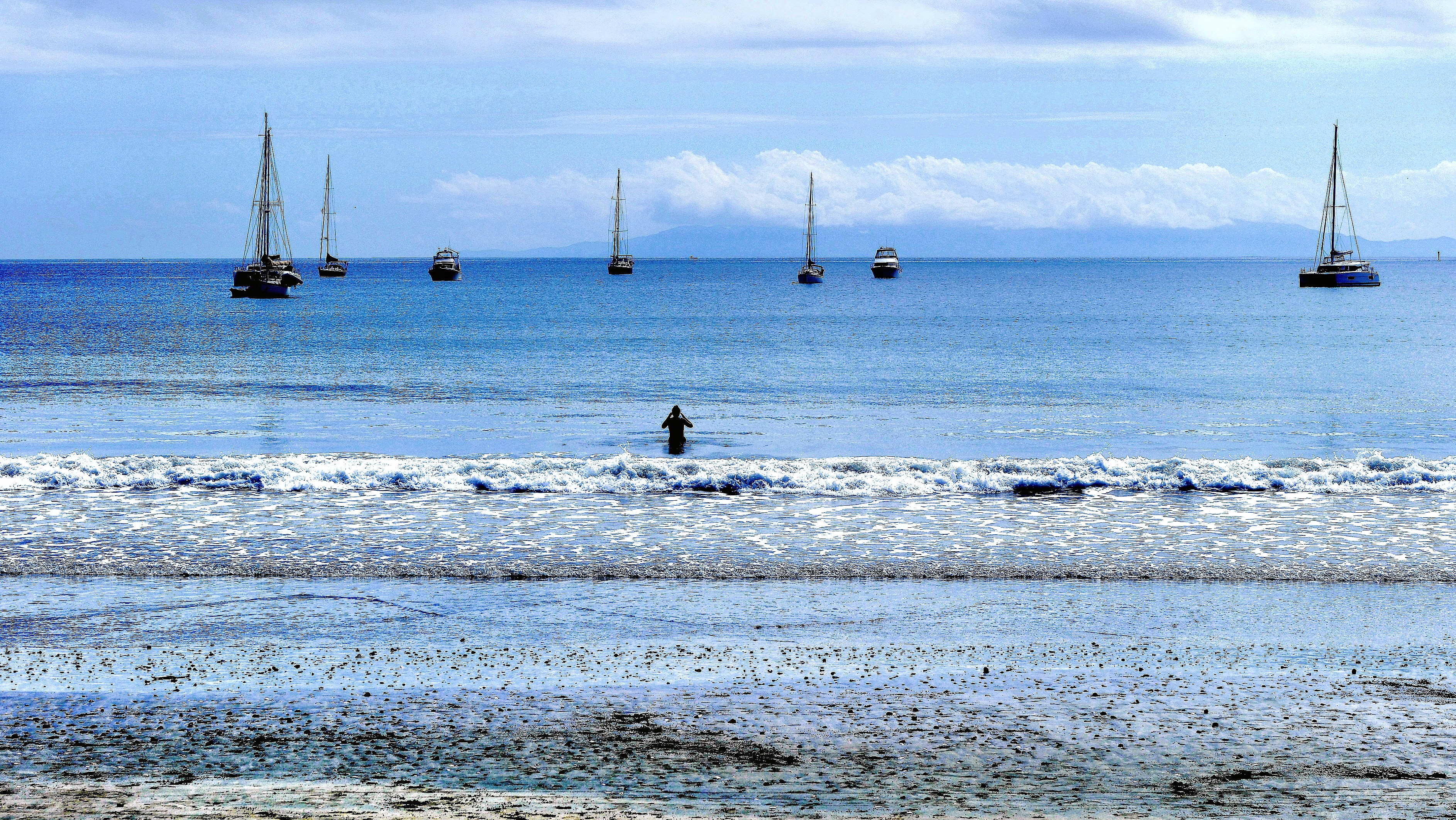 A person standing in shallow water at the beach, with several sailing boats anchored in the calm sea in the background under a partly cloudy sky.