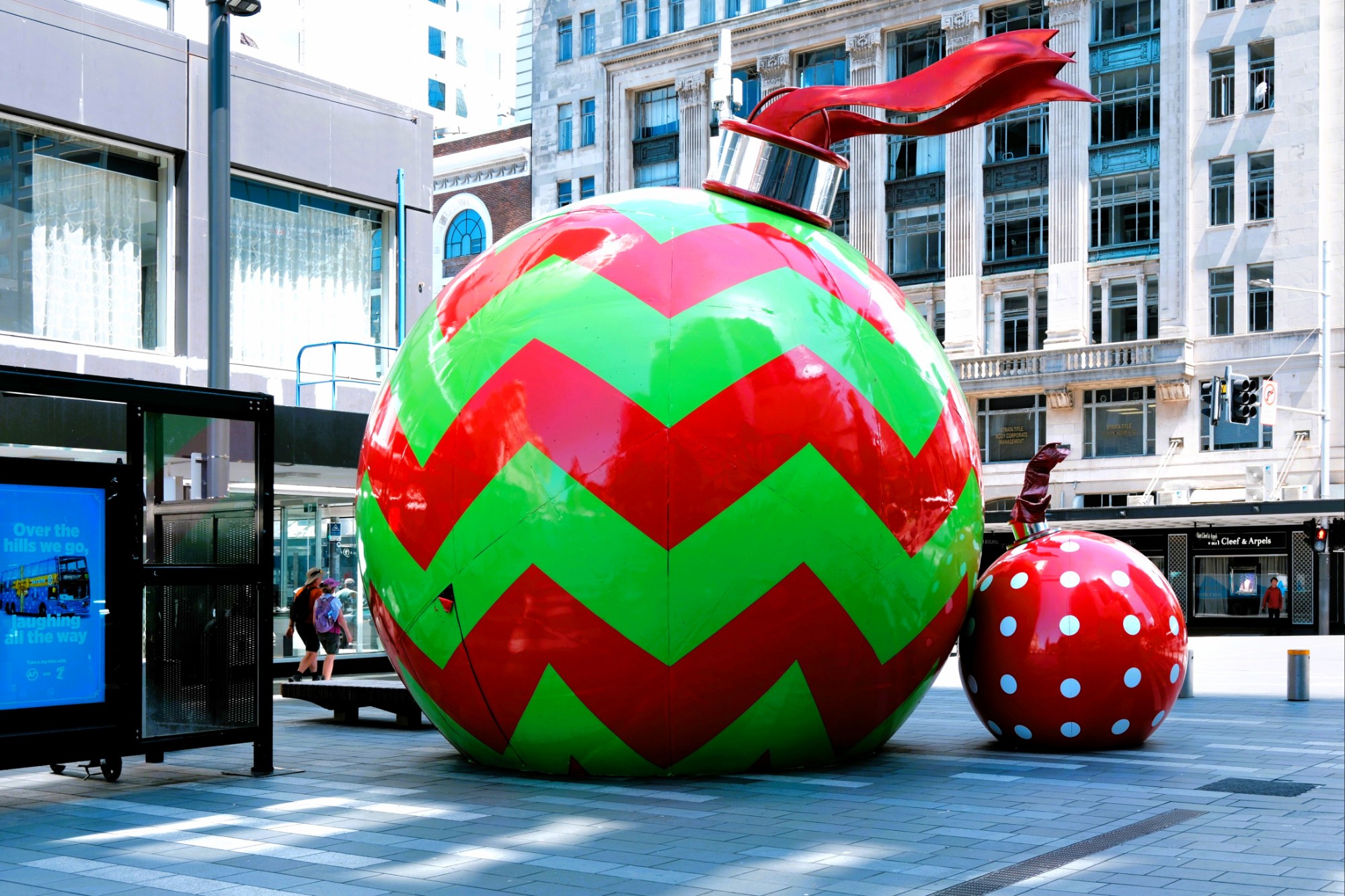 Large festive ornaments in a city plaza, including a green and red chevron-patterned ornament and a smaller red ornament with white polka dots.