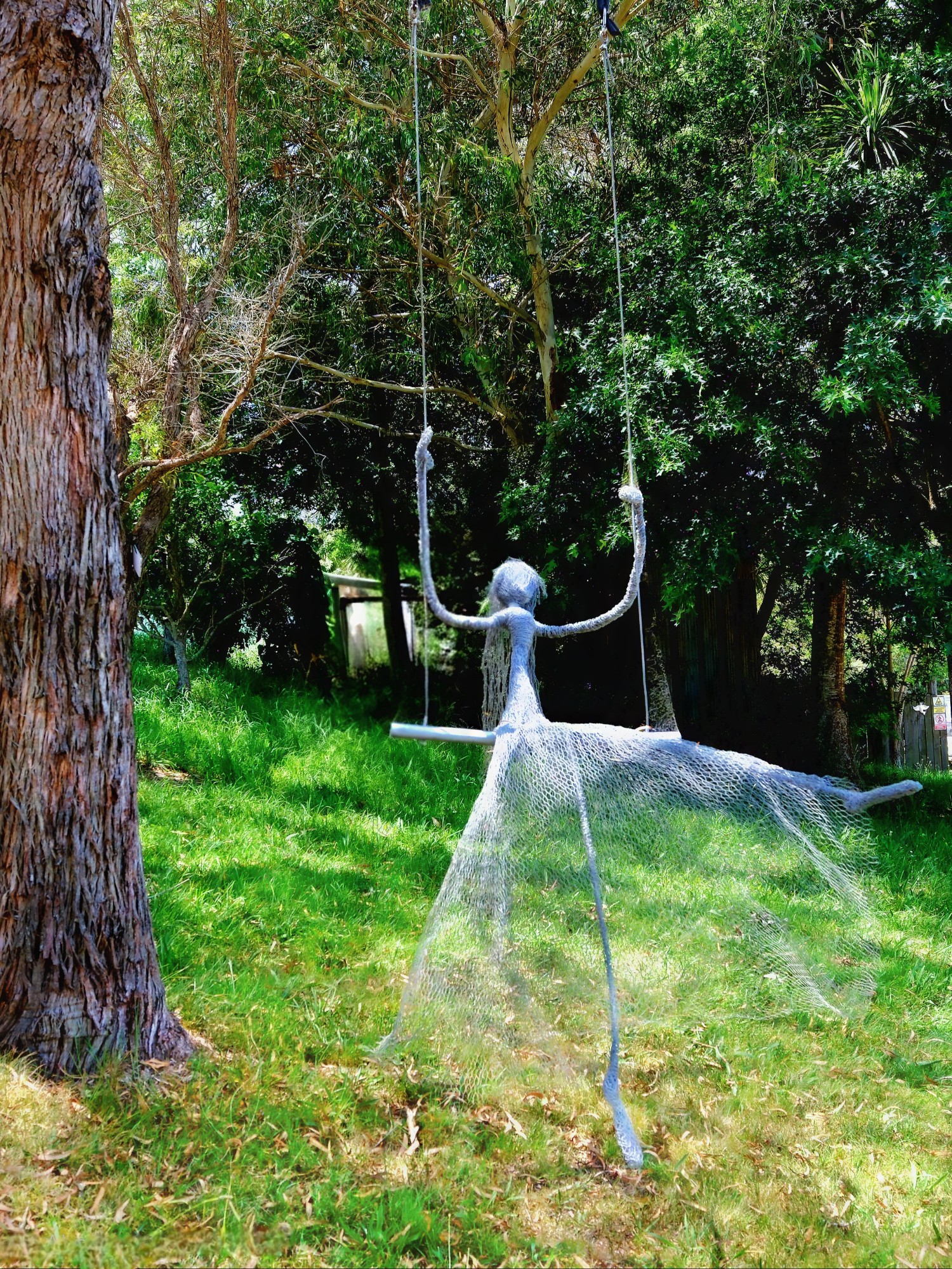 A wire sculpture of a figure on a swing in a grassy area surrounded by trees.