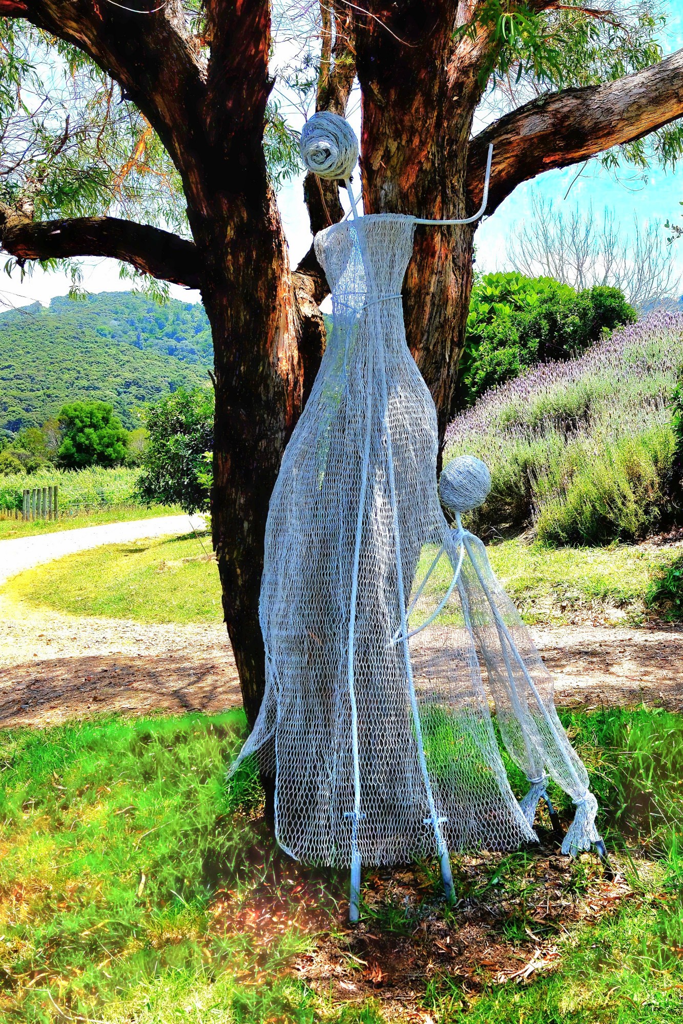 A large wire sculpture of a woman and child standing under a tree in a green field, with a pathway and mountains in the background.